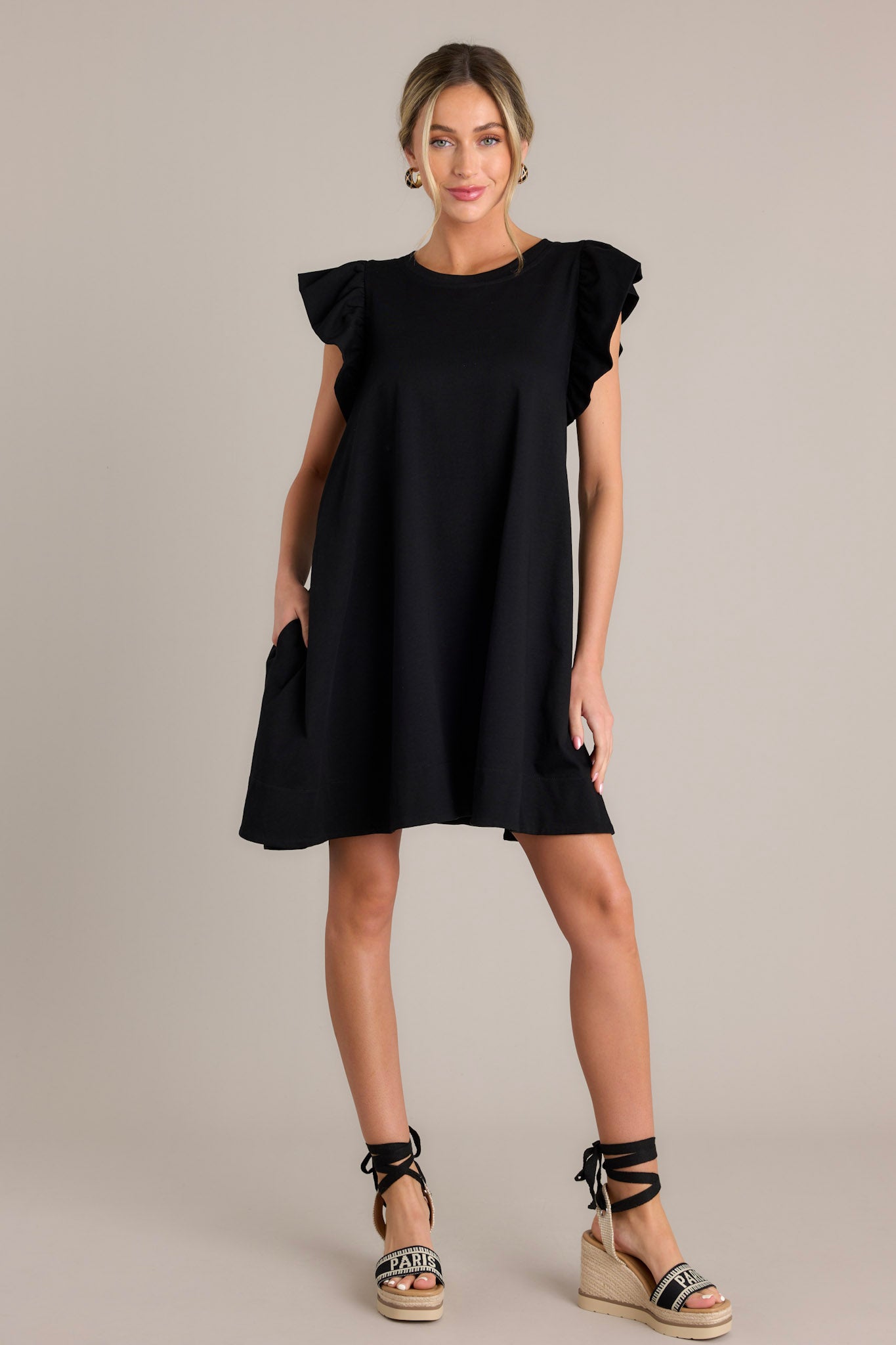 This black mini dress features a crew neckline, a self-tie drawstring waist feature in the back of the dress, functional hip pockets, and ruffled short sleeves.
