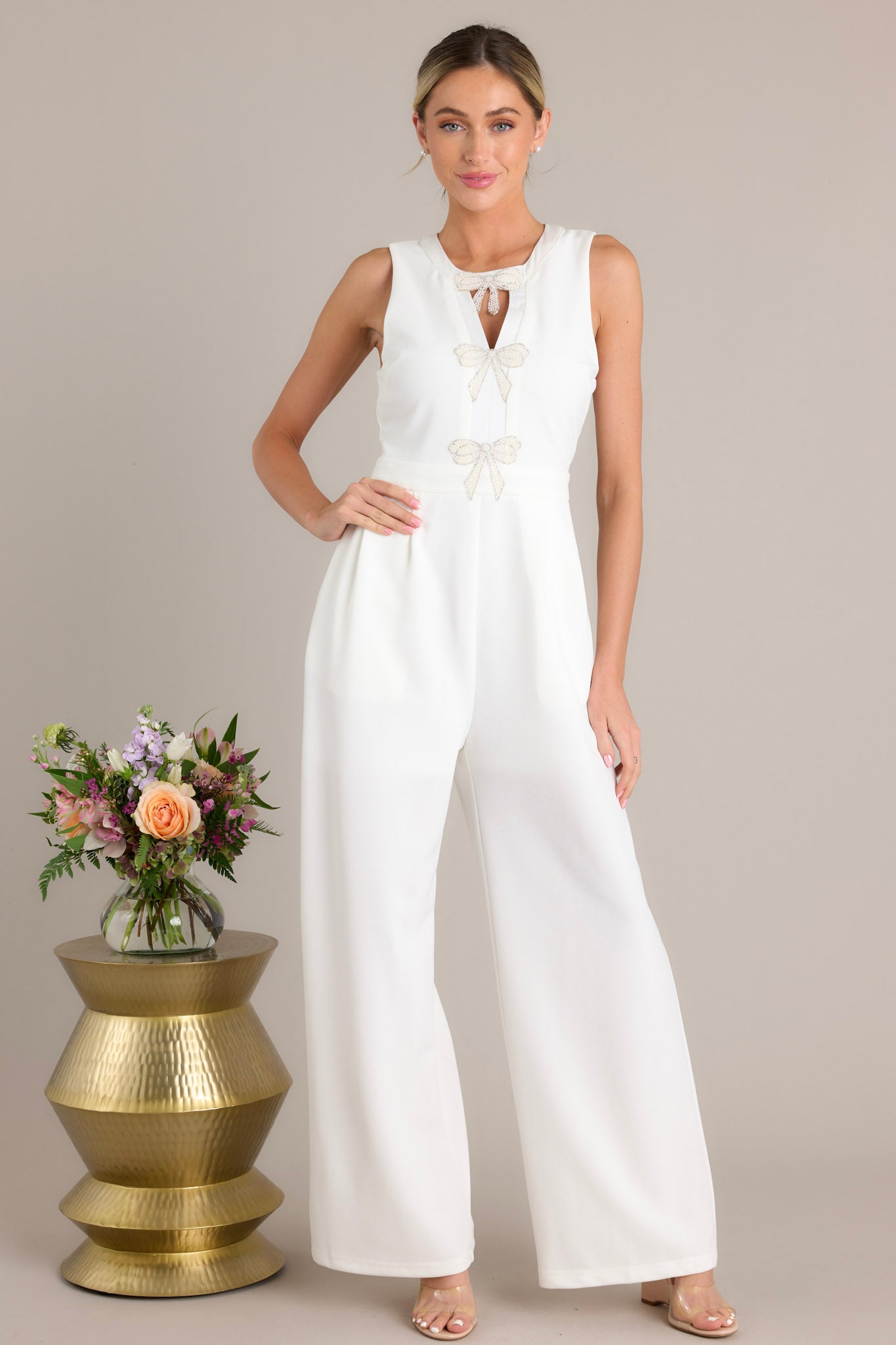 Full body front view of this white jumpsuit that features a round neckline, a discrete back zipper, three ornate bows embellished with faux ivory pearls and rhinestones, a chest cutout, a thick waistband, subtle pleats, functional hip pockets, and a wide leg design.
