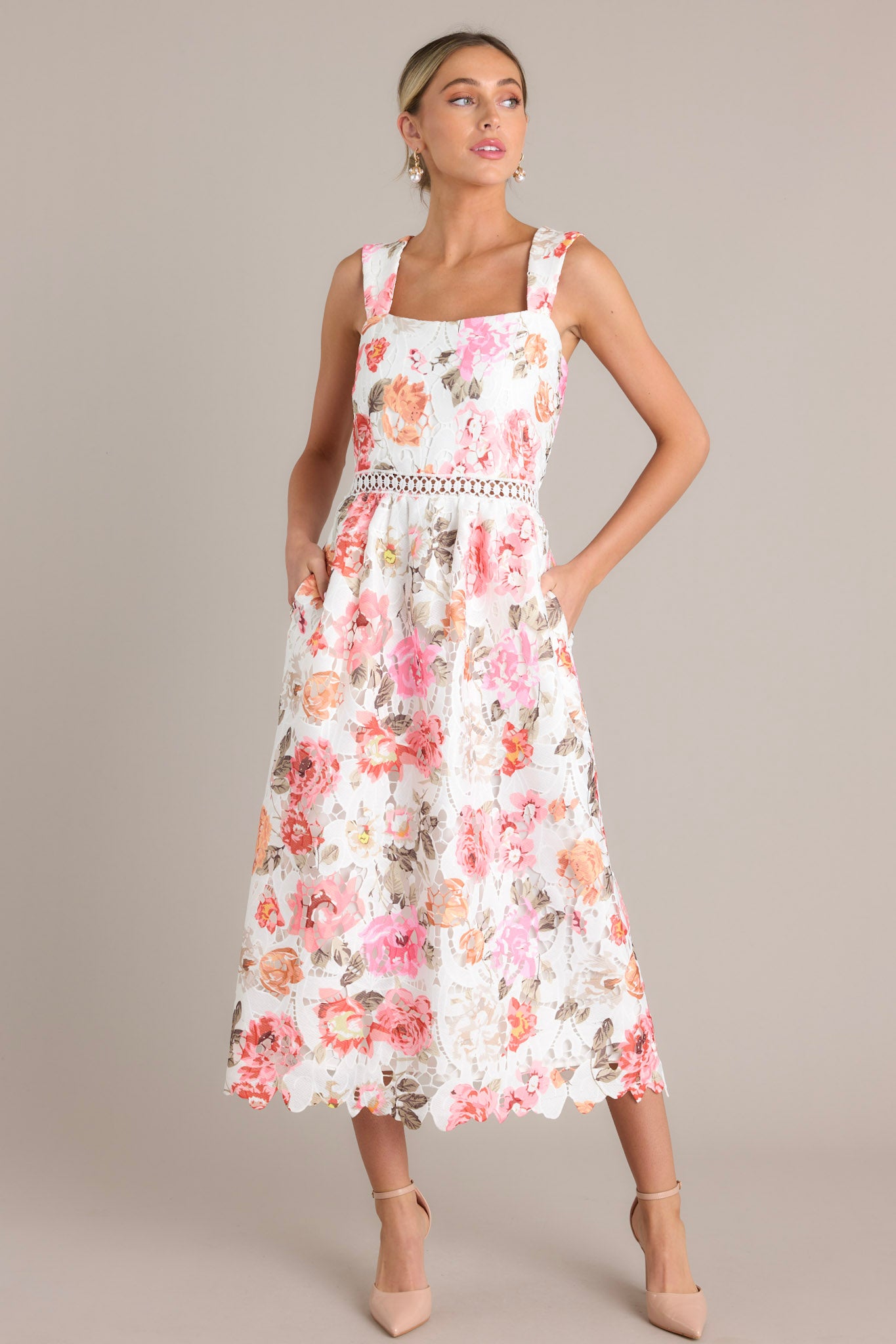 This white multi floral midi dress features a square neckline, a fitted bodice, a crocheted cinched waist, and functional pockets, complemented by a full lace mid-length skirt.