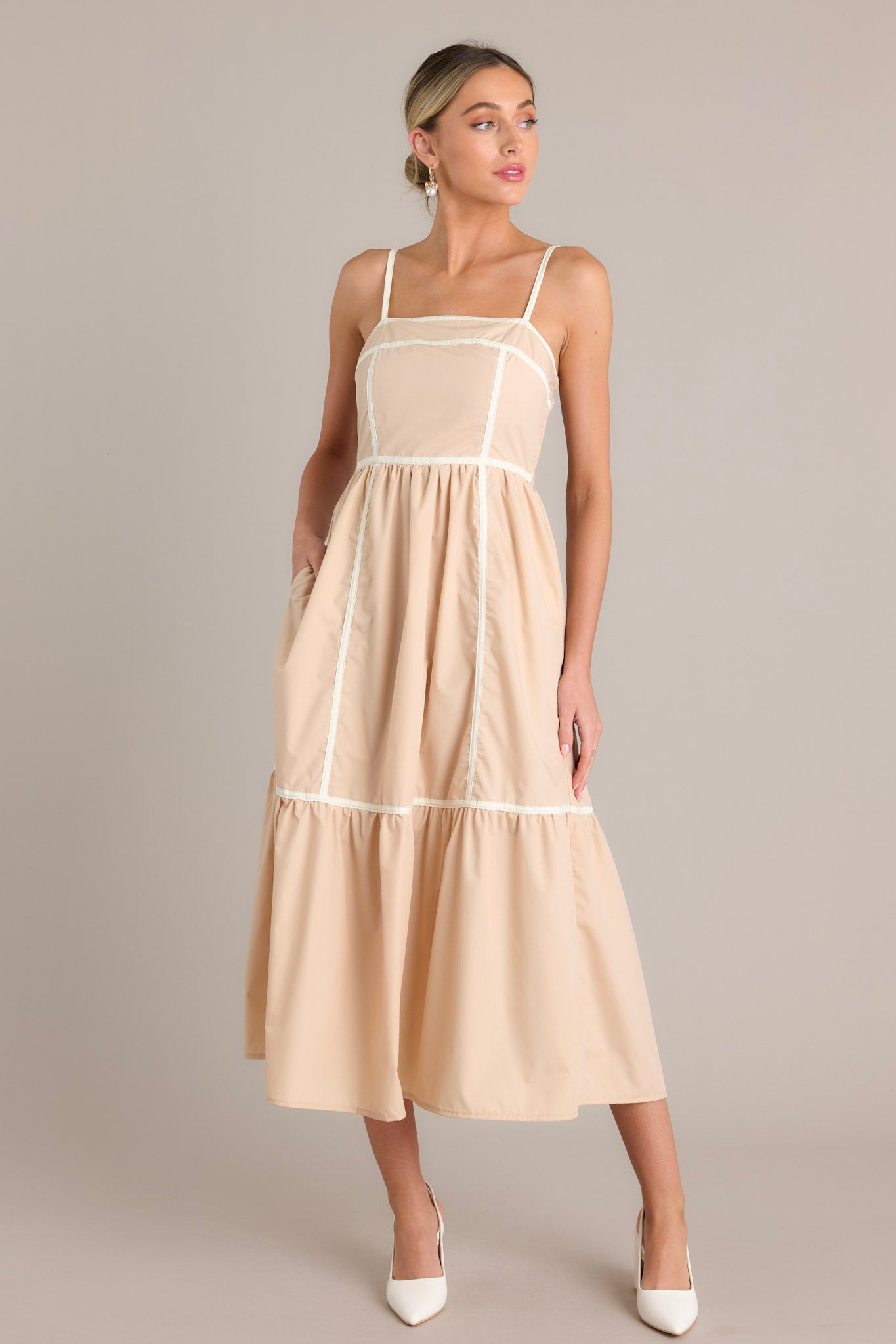 Full length view of a beige maxi dress with a square neckline, thin adjustable straps, a fully smocked back, functional hip pockets, and thick contrasting seams throughout