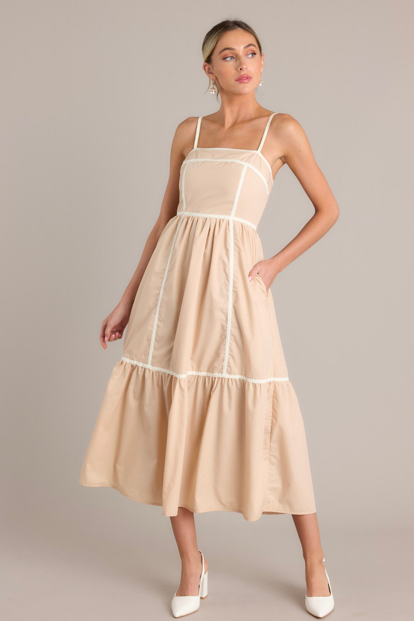 Front angled view of a beige maxi dress featuring a square neckline, thin adjustable straps, a fully smocked back, functional hip pockets, and thick contrasting seams throughout