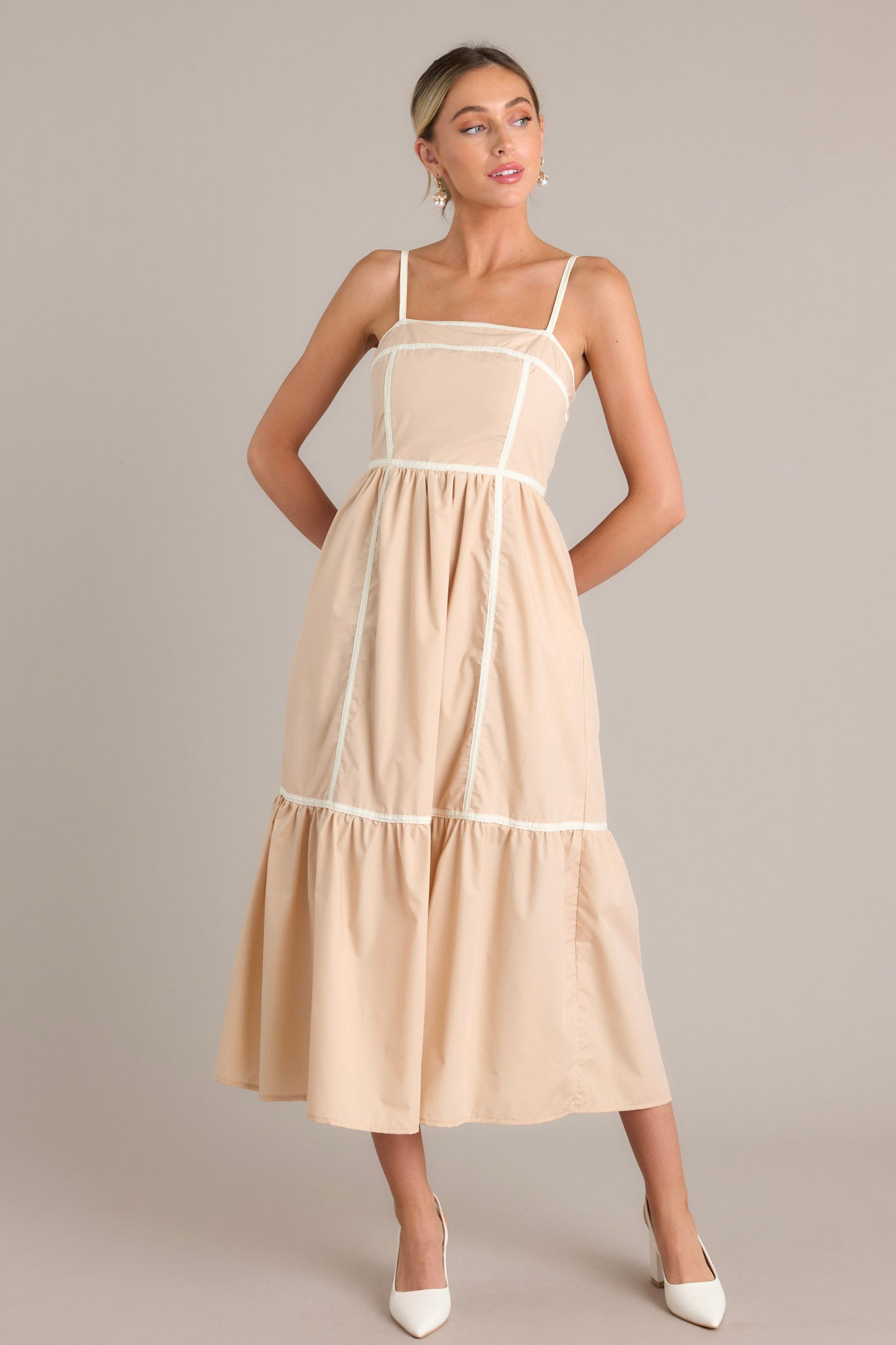Front view of a beige maxi dress featuring a square neckline, thin adjustable straps, functional hip pockets, and thick contrasting seams throughout.