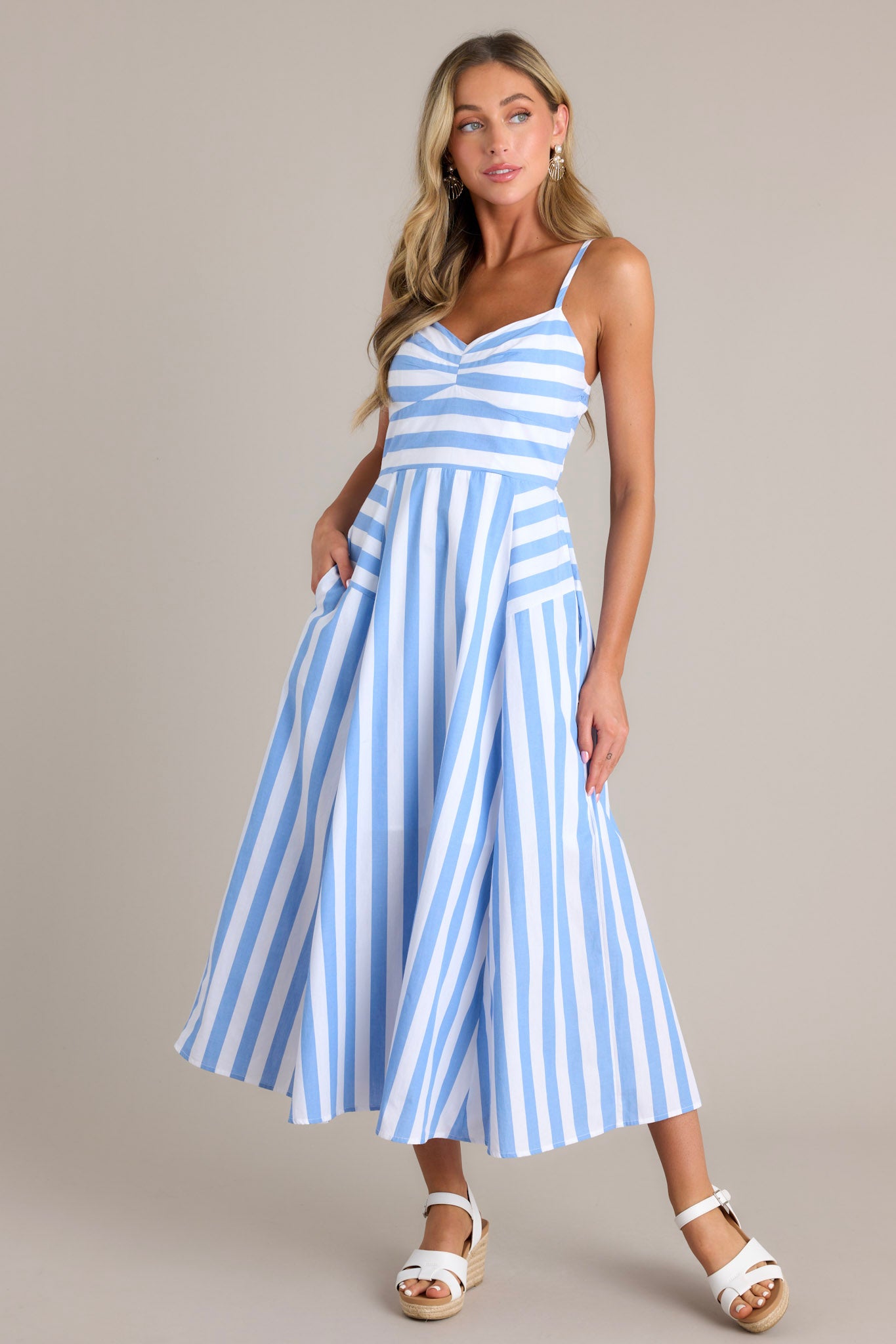 This blue stripe midi dress features a sweetheart neckline, thin adjustable straps, a fully smocked back, functional hip pockets, and a unique striped pattern.