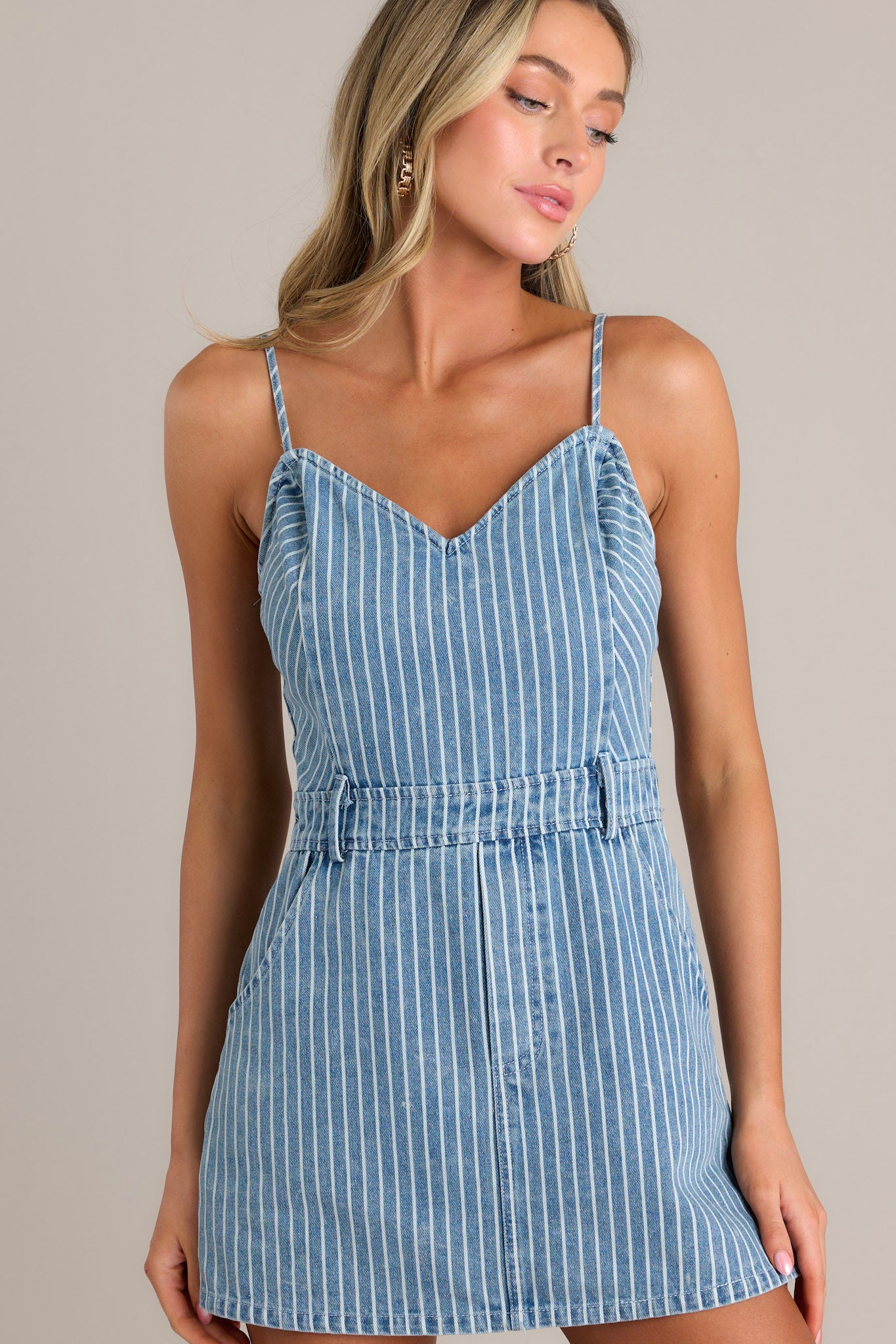 Front angled view of a denim romper featuring a v-neckline, thin adjustable straps, a thick waistline, belt loops, functional hip & back pockets, and hidden shorts