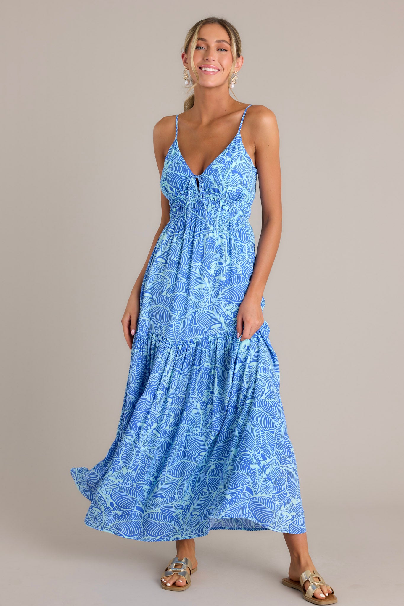 This blue maxi dress features a v-neckline, thin adjustable straps, a self-tie bust feature, an elastic waistband, a single tier, and a flowing silhouette.