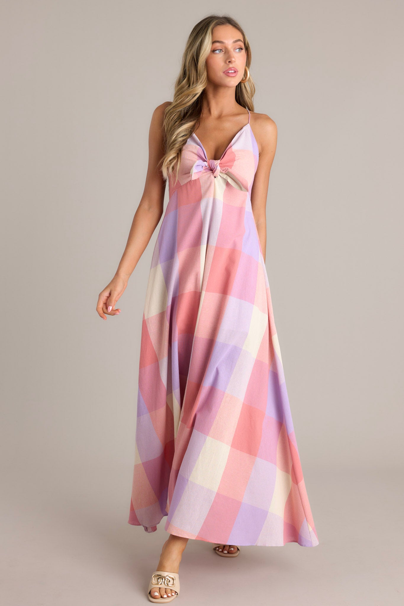 Full length view of a pink maxi dress featuring a v-neckline, a self-tie bust feature, thin self-tie straps that cross in the back, an open back, a discrete back zipper, a large plaid pattern, and a 22.5" front slit