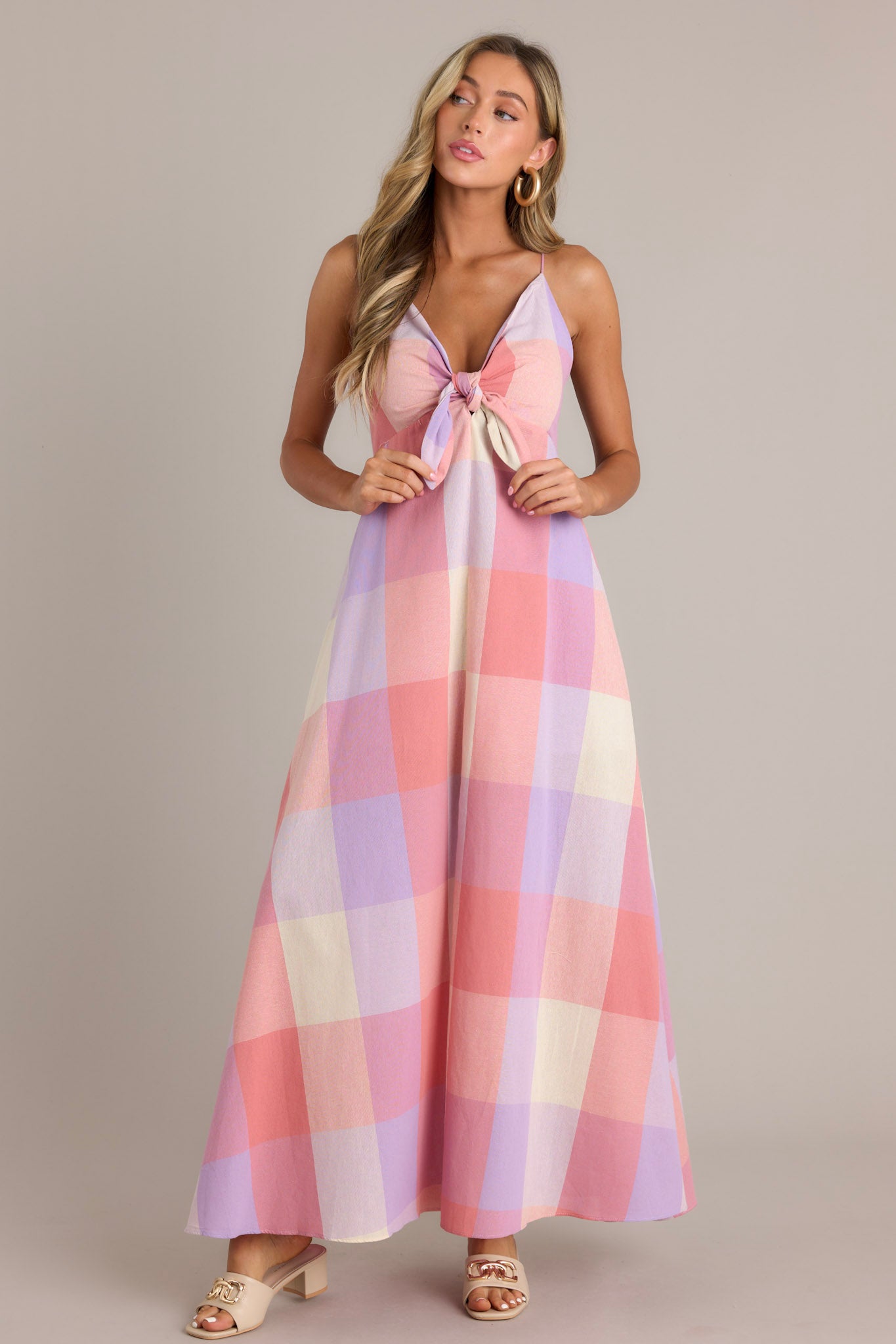 Front view of a pink maxi dress featuring a v-neckline, a self-tie bust feature, thin self-tie straps that cross in the back, an open back, a discrete back zipper, a large plaid pattern, and a 22.5" front slit