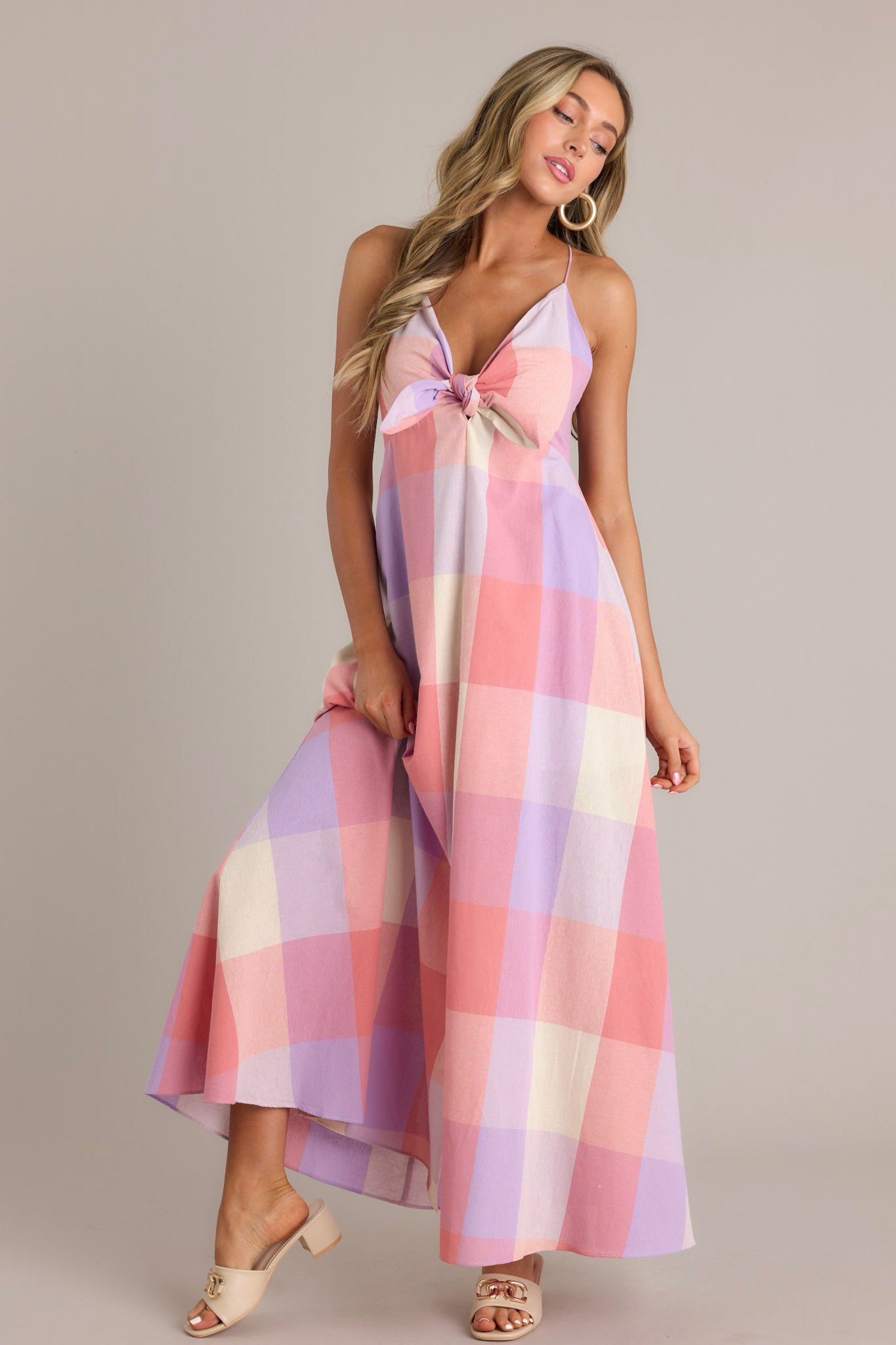 Front angled view of a pink maxi dress featuring a v-neckline, a self-tie bust feature, thin self-tie straps that cross in the back, an open back, a discrete back zipper, a large plaid pattern, and a 22.5" front slit