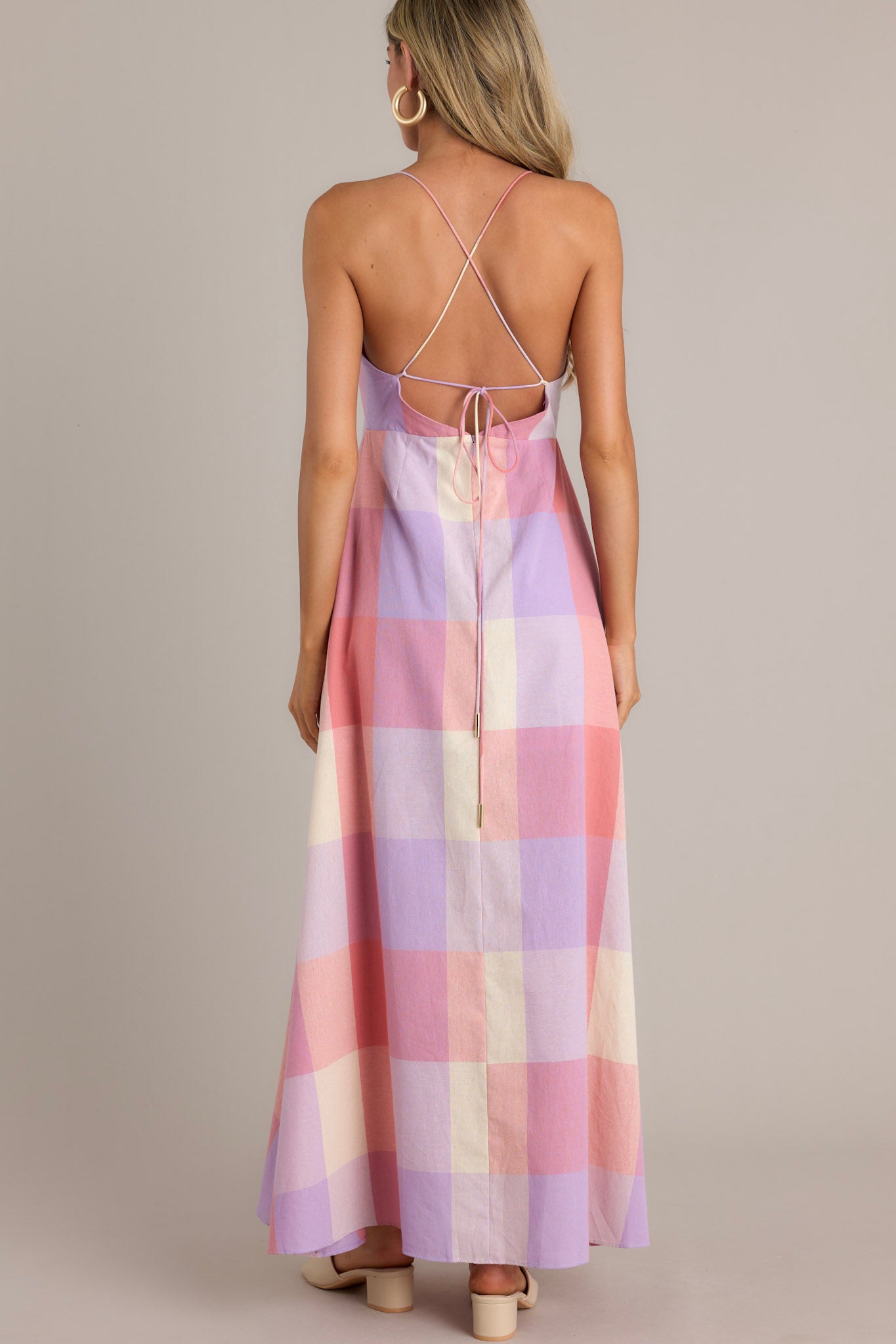 Back view of a pink maxi dress featuring an open back with thin self-tie straps that cross, a discrete back zipper, and a flowing plaid pattern.