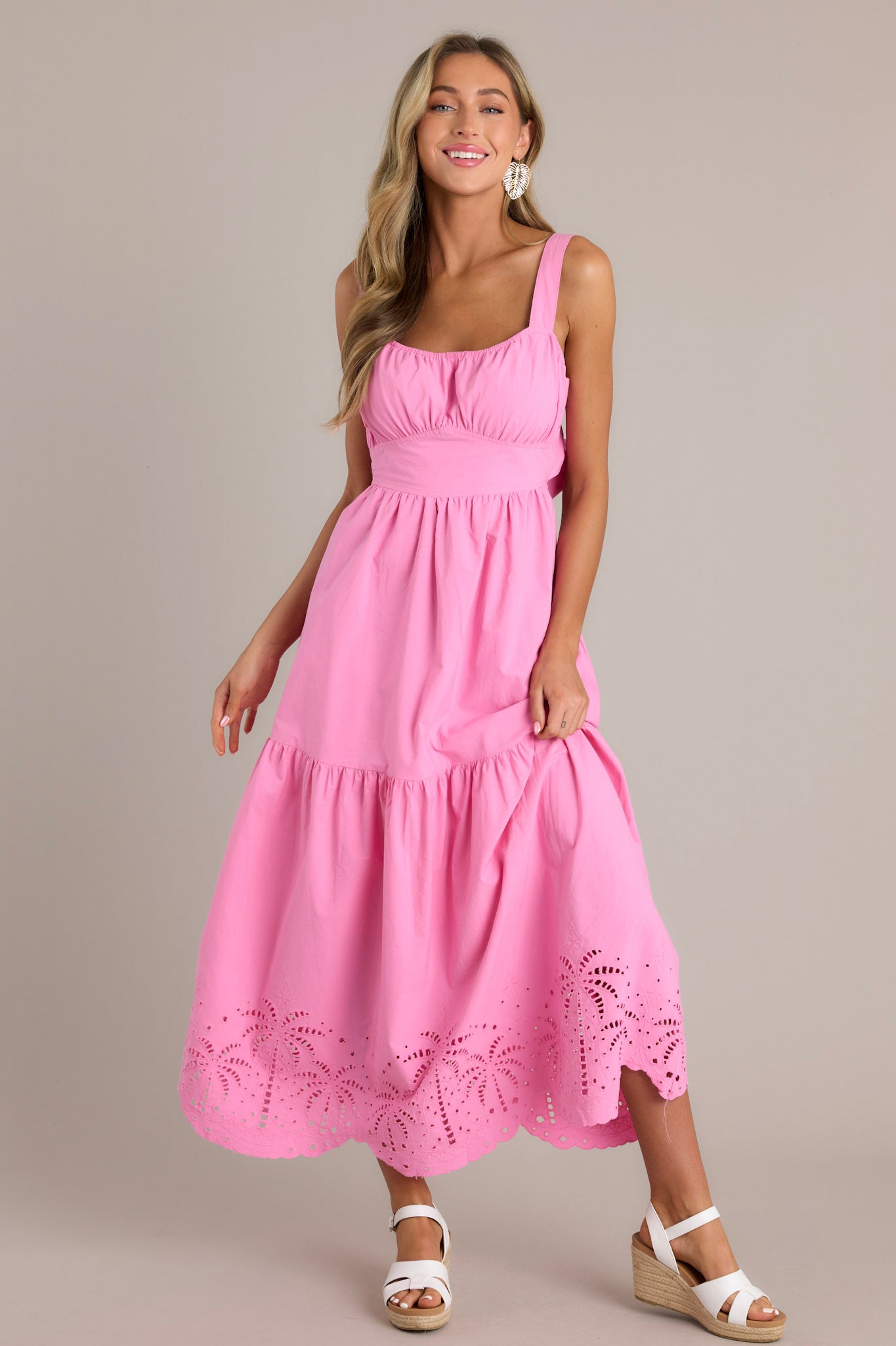 Full length view of a model wearing a pink midi dress with an elastic square neckline, thick adjustable straps, thick waistband with a self-tie back feature, open lower back, single tier, palm eyelet detailing, and scalloped hemline.