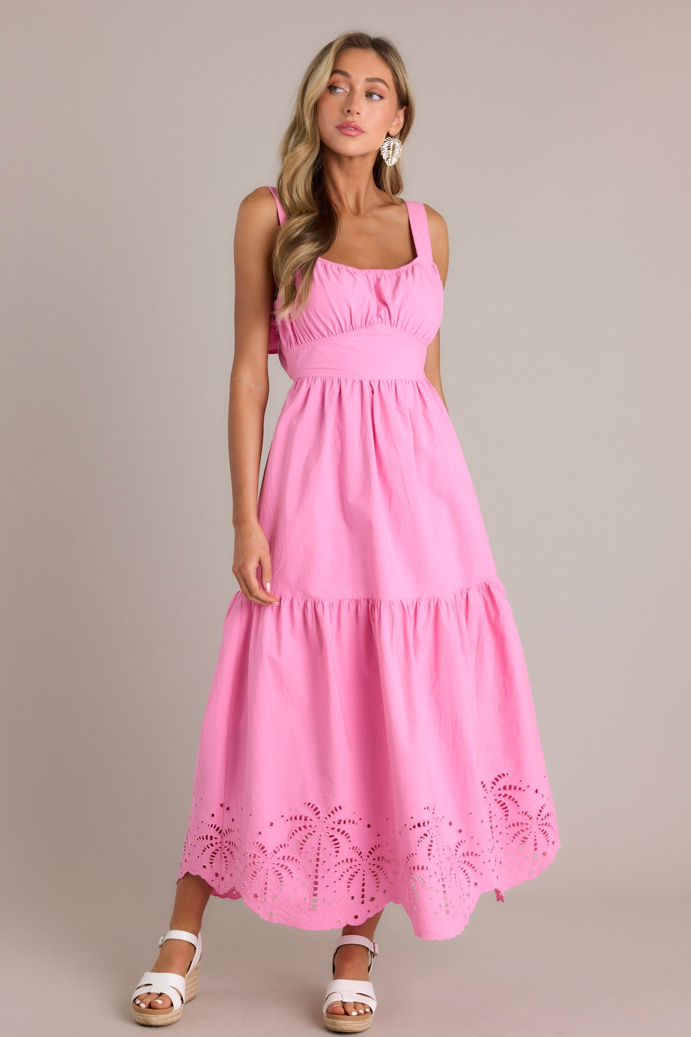 Front angled view of a pink midi dress featuring an elastic square neckline, thick adjustable straps, a thick waistband with a self-tie back feature, an open lower back, a single tier, palm eyelet detailing, and a scalloped hemline