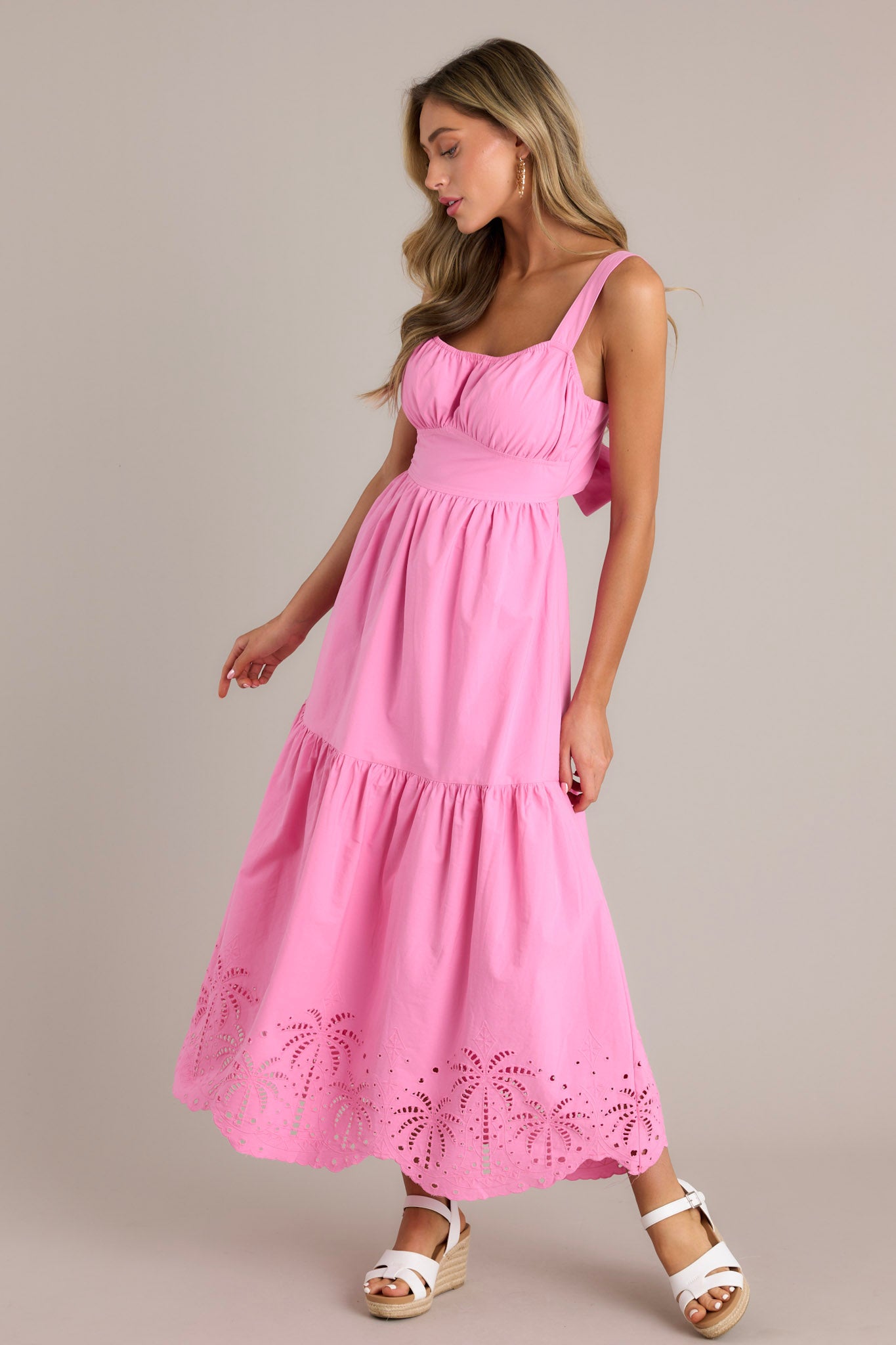 Action shot of a pink midi dress displaying the flow and movement of the fabric, highlighting the elastic square neckline, thick adjustable straps, thick waistband with a self-tie back feature, open lower back, and the palm eyelet detailing on the scalloped hemline.