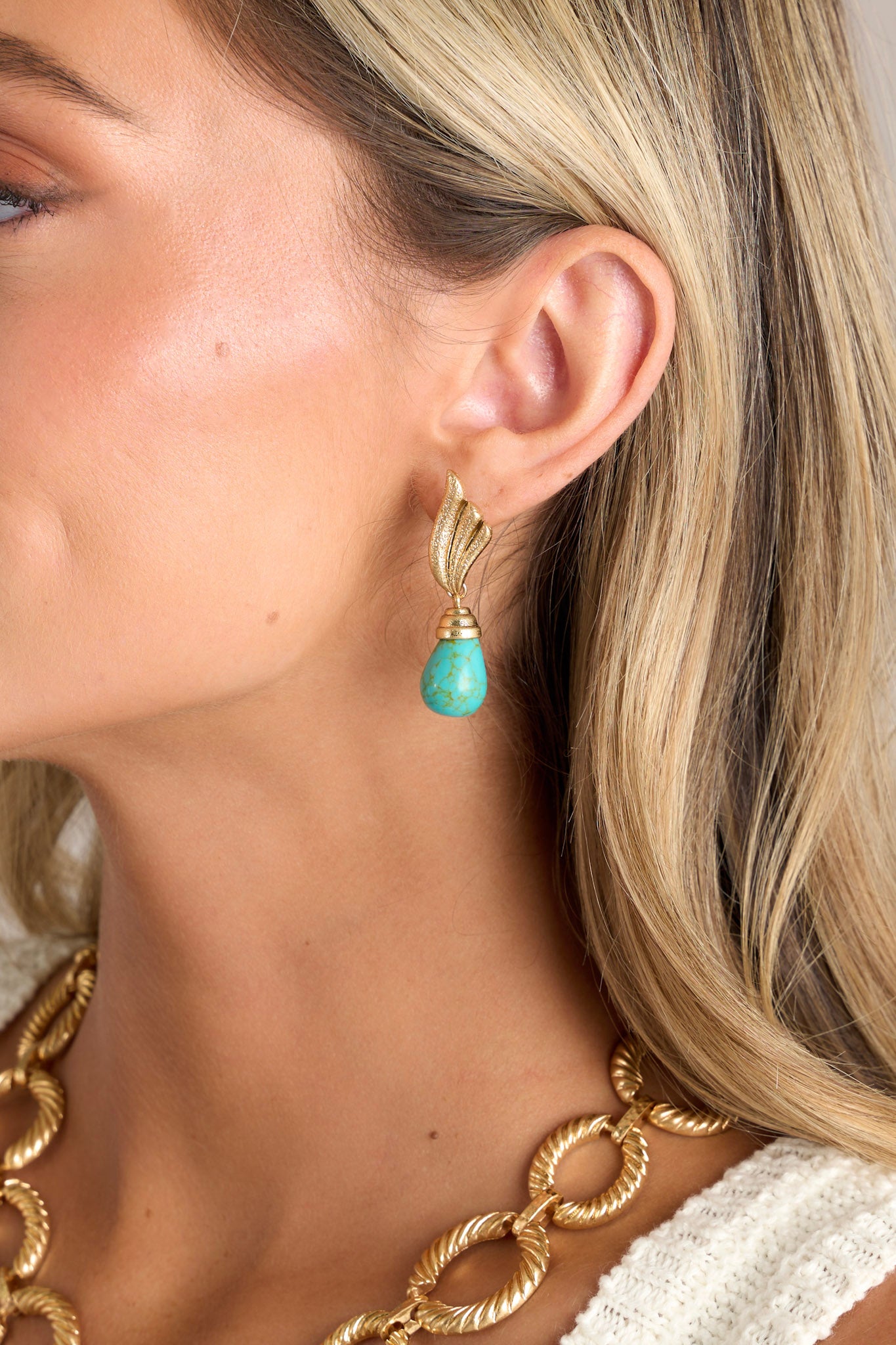 Close-up view of these gold earrings that feature textured winged studs, a turquoise stone drop pendant, and secure post backings.