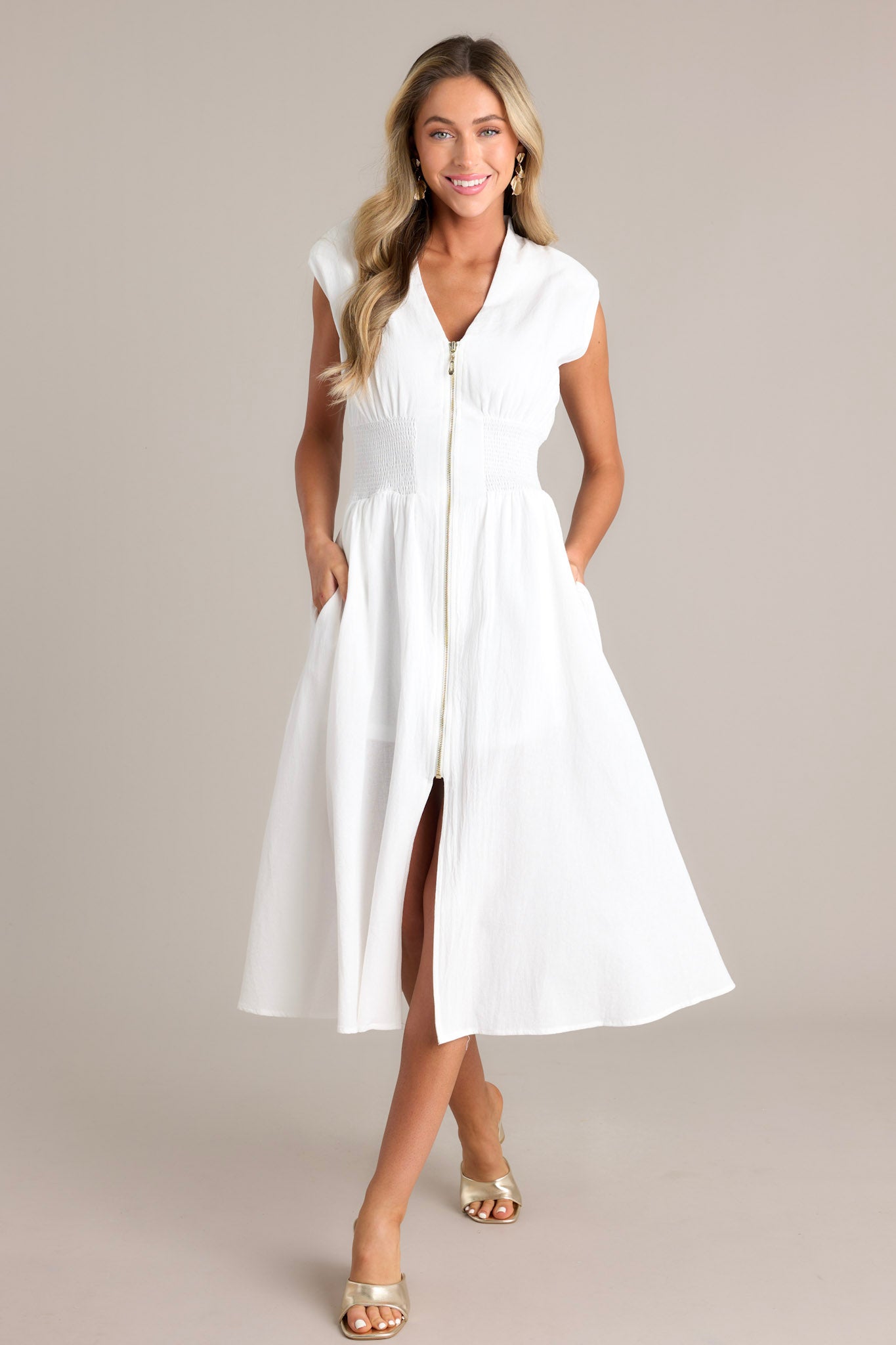 Full length view of a white midi dress featuring a deep v-neckline, shoulder padding, a functional zipper front, a fully smocked waist, functional pockets, and a front slit