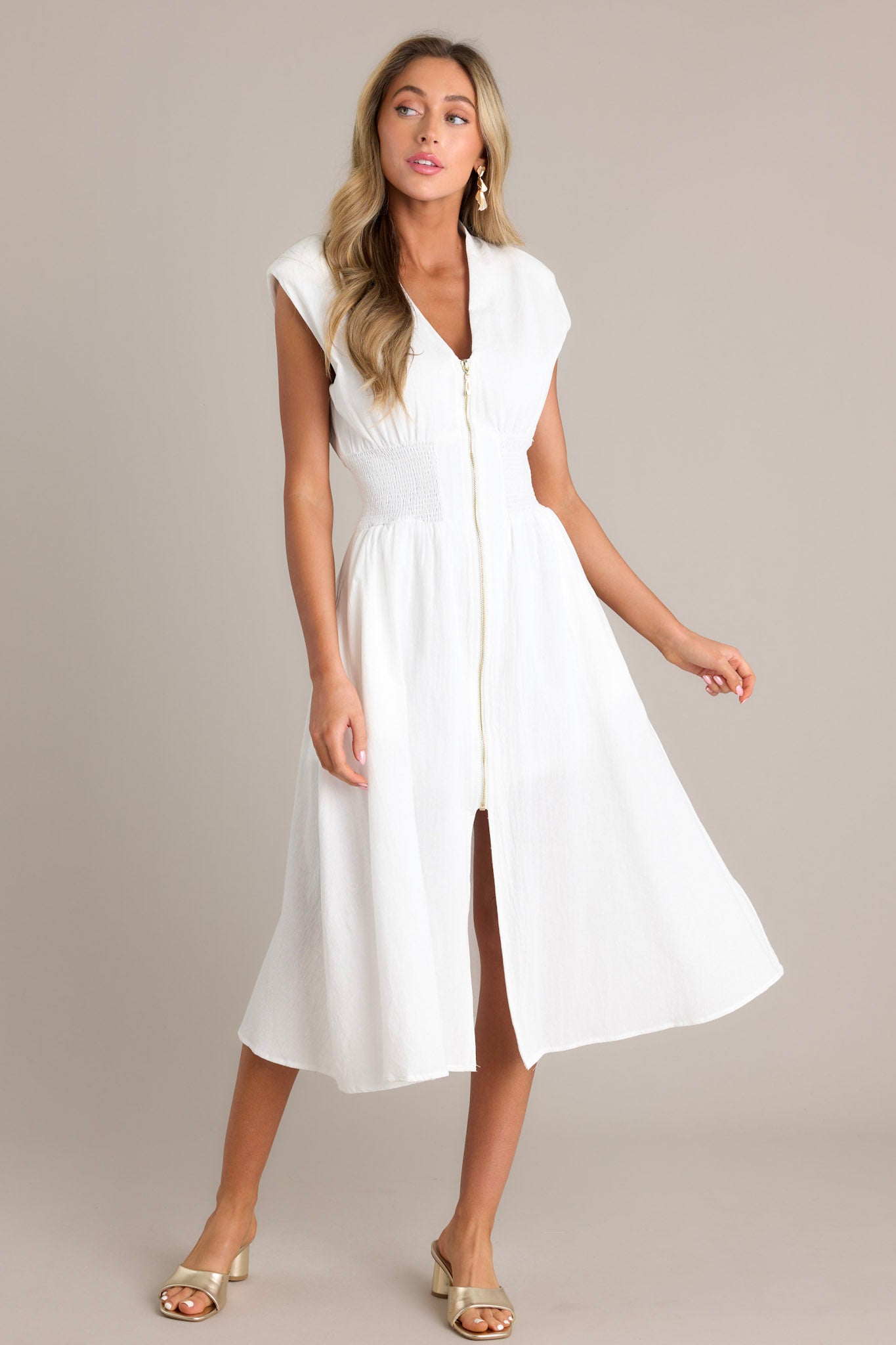 Front angled view of a white midi dress featuring a deep v-neckline, shoulder padding, a functional zipper front, a fully smocked waist, functional pockets, and a front slit