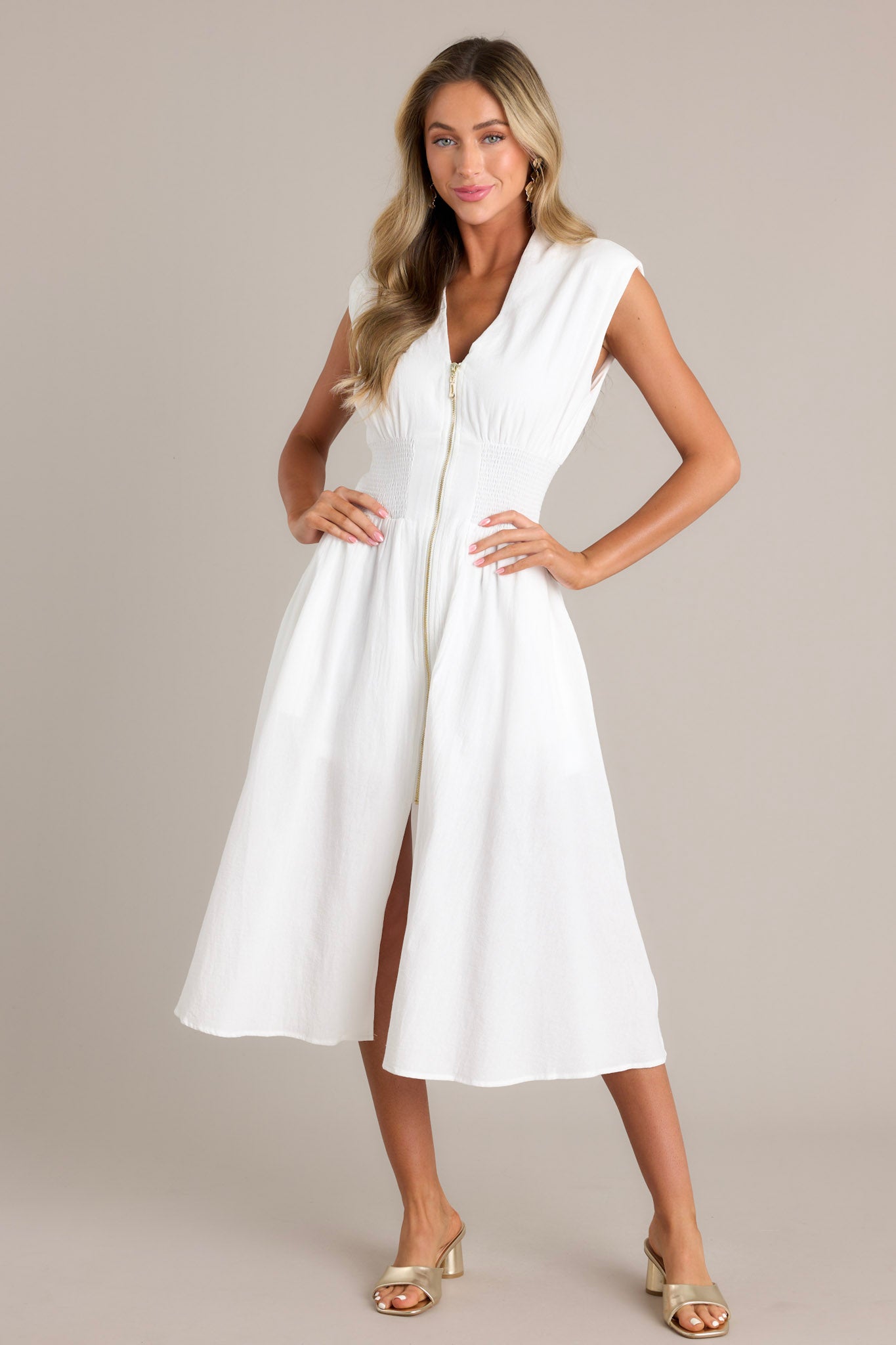 Front view of a white midi dress featuring a deep v-neckline, shoulder padding, a functional zipper front, a fully smocked waist, functional pockets, and a front slit.