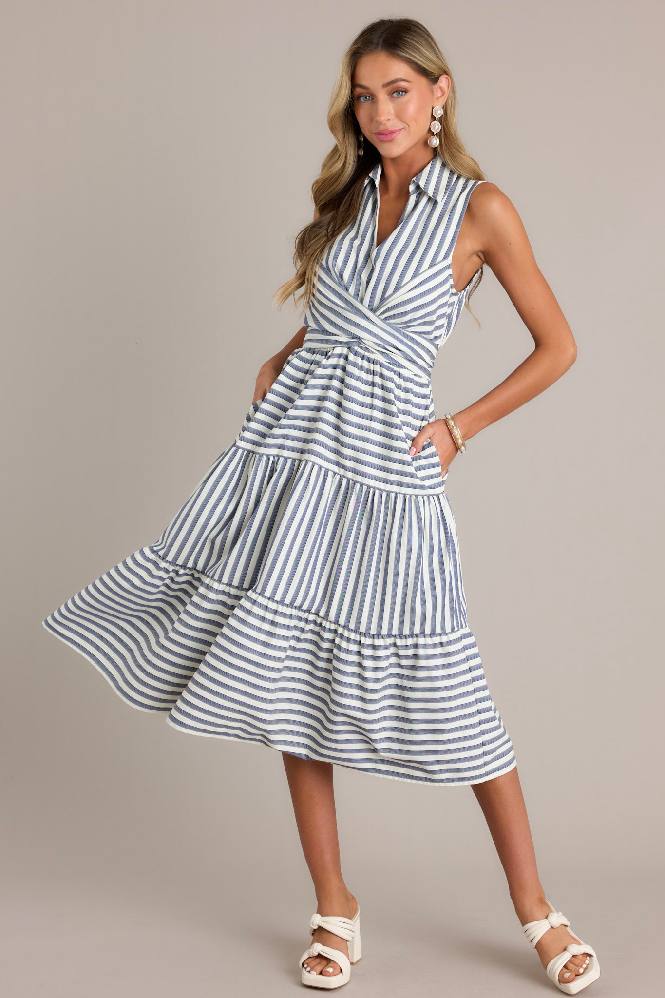 Full body front view of this navy stripe midi dress that features a collared v-neckline, a self-tie waist feature that can be in the front or back, an elastic waist insert, functional hip pockets, and a sleeveless design.