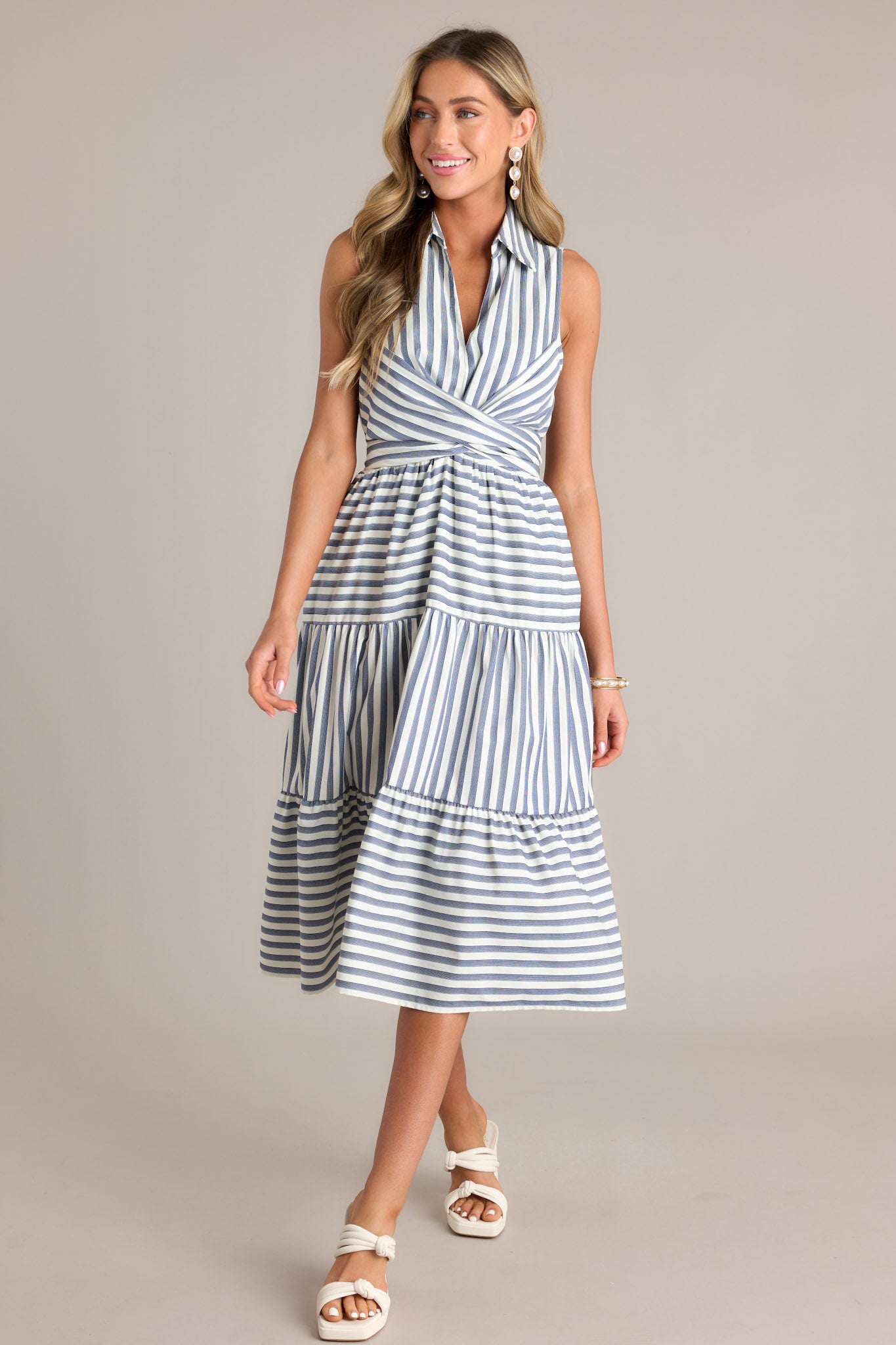 Full body view of this navy stripe midi dress that features a collared v-neckline, a self-tie waist feature that can be in the front or back, an elastic waist insert, functional hip pockets, and a sleeveless design.