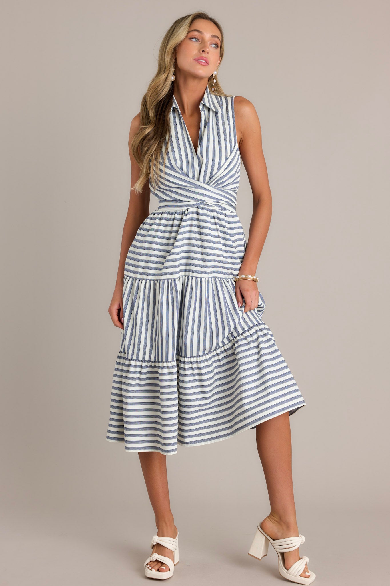 This navy stripe midi dress features a collared v-neckline, a self-tie waist feature that can be in the front or back, an elastic waist insert, functional hip pockets, and a sleeveless design.