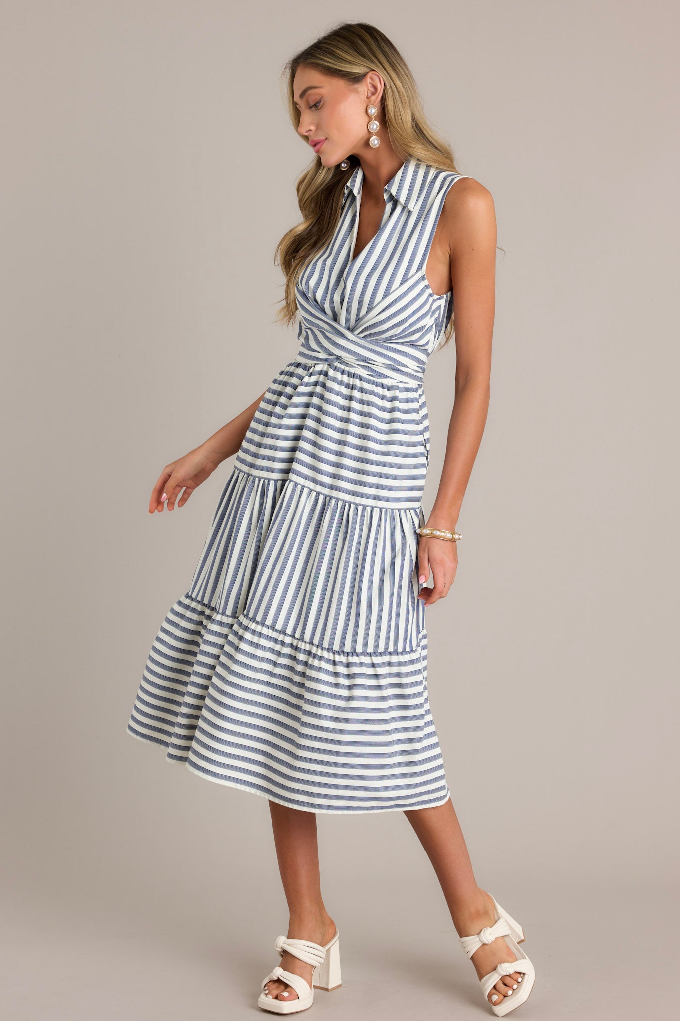 Side view of this navy stripe midi dress that features a collared v-neckline, a self-tie waist feature that can be in the front or back, an elastic waist insert, functional hip pockets, and a sleeveless design.