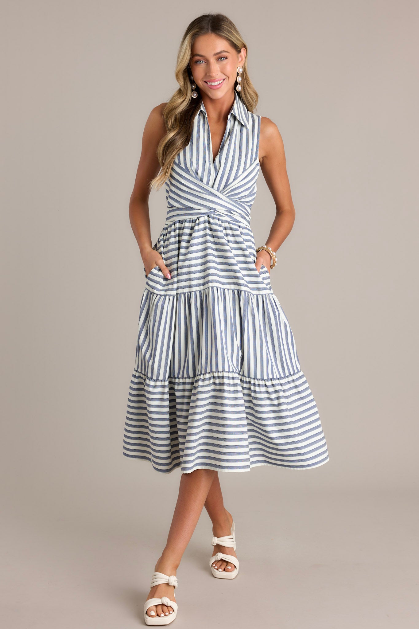 Front view of this navy stripe midi dress that features a collared v-neckline, a self-tie waist feature that can be in the front or back, an elastic waist insert, functional hip pockets, and a sleeveless design.