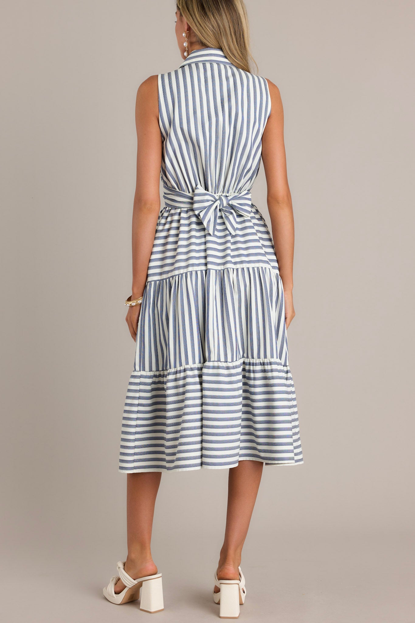 Back view of this navy stripe midi dress that features a collared v-neckline, a self-tie waist feature that can be in the front or back, an elastic waist insert, functional hip pockets, and a sleeveless design.