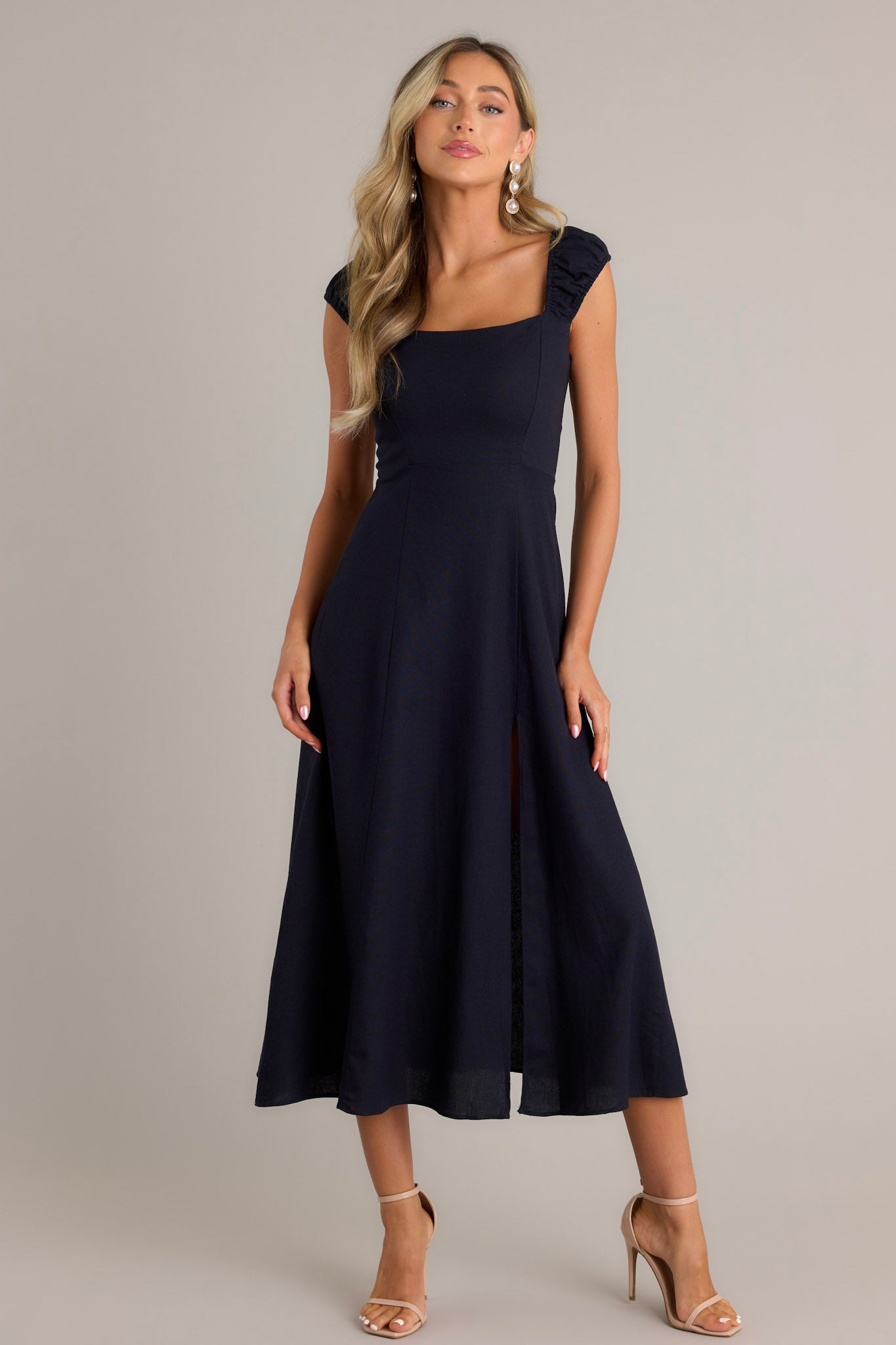 Full length view of a navy midi dress with a square neckline, smocked sleeves, smocked back section, hidden zipper, slit up the leg, and a flowy skirt.