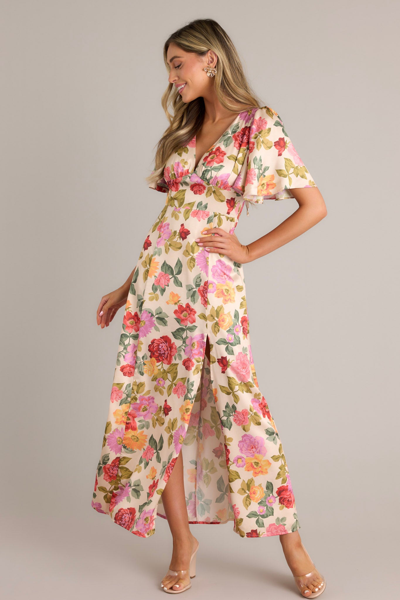 This beige multi floral maxi dress features a deep V-neckline, flutter sleeves, a warm floral print, a fitted waist, an A-line skirt, and a front slit.