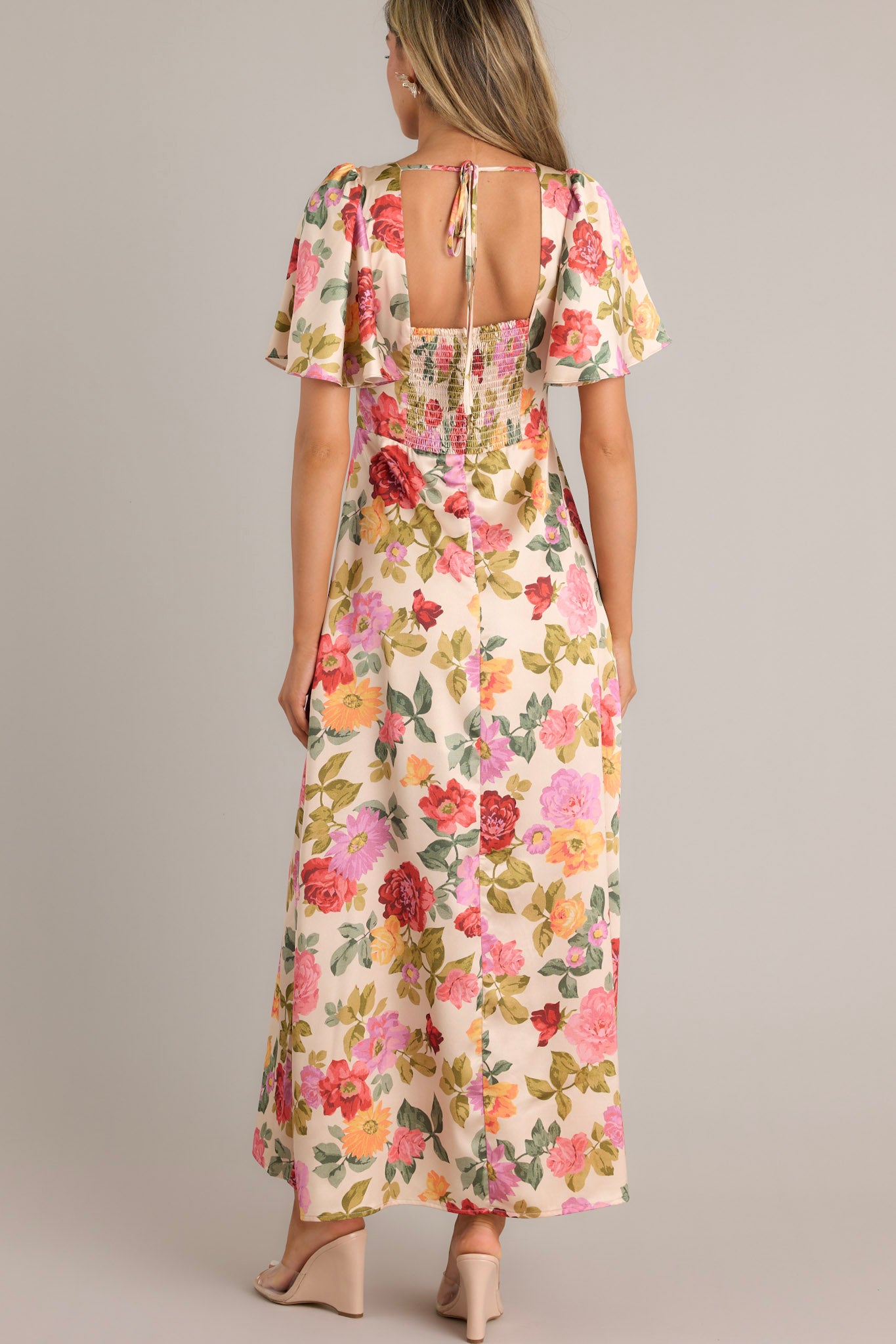 Back view of this beige floral maxi dress featuring a deep V-neckline, flutter sleeves, fitted waist, and A-line skirt with a warm floral print.