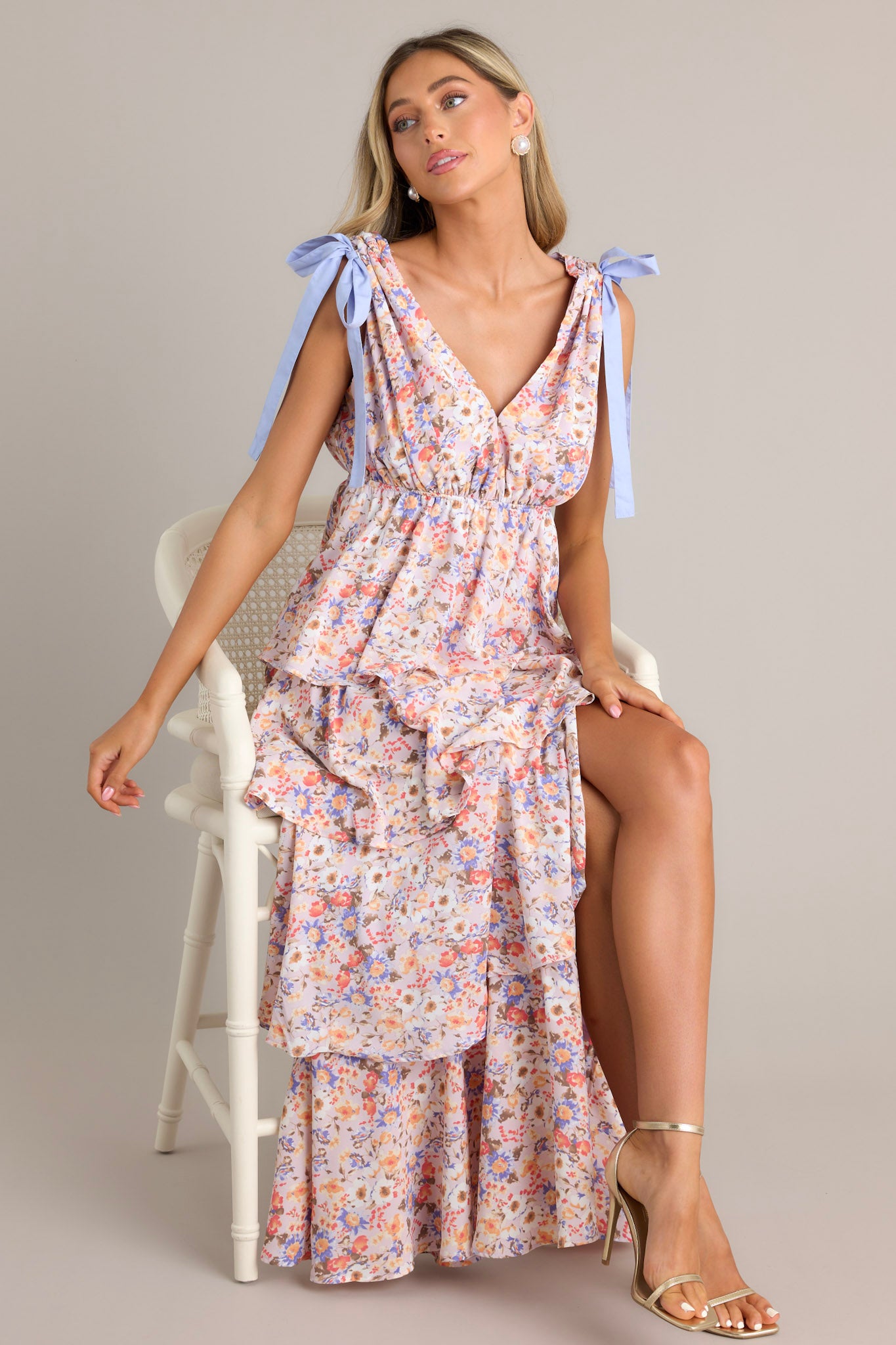 Seated view of this lavender floral dress featuring an empire waist, a V neckline, self ties at the top of the thick sleeveless gathered straps, an elastic waist, a slit up the front, and tier layers down the skirt.