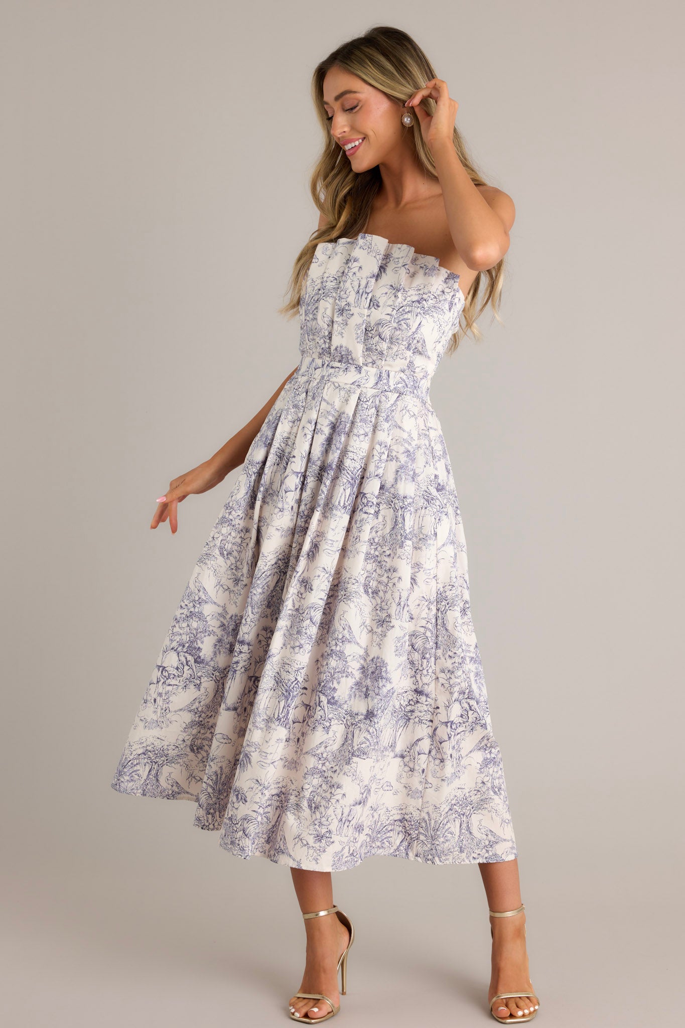 Movement shot of this blue toile strapless midi dress with a structured, pleated bodice, functional pockets, and a smocked back.