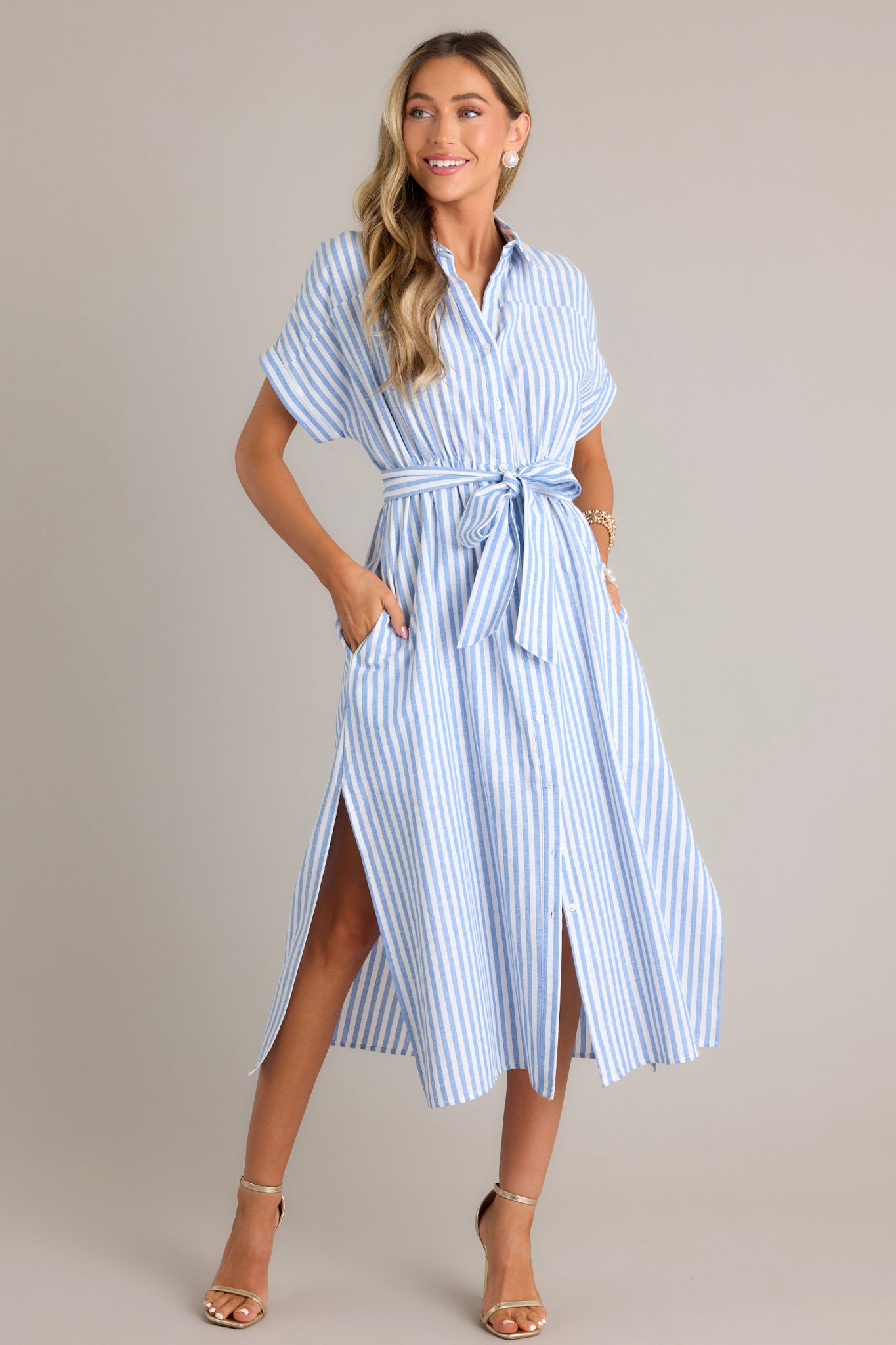 This white and blue dress features a collared neckline, a full button front, a fitted waist, a self-tie waist belt, functional pockets, two side slits, and folded short sleeves.