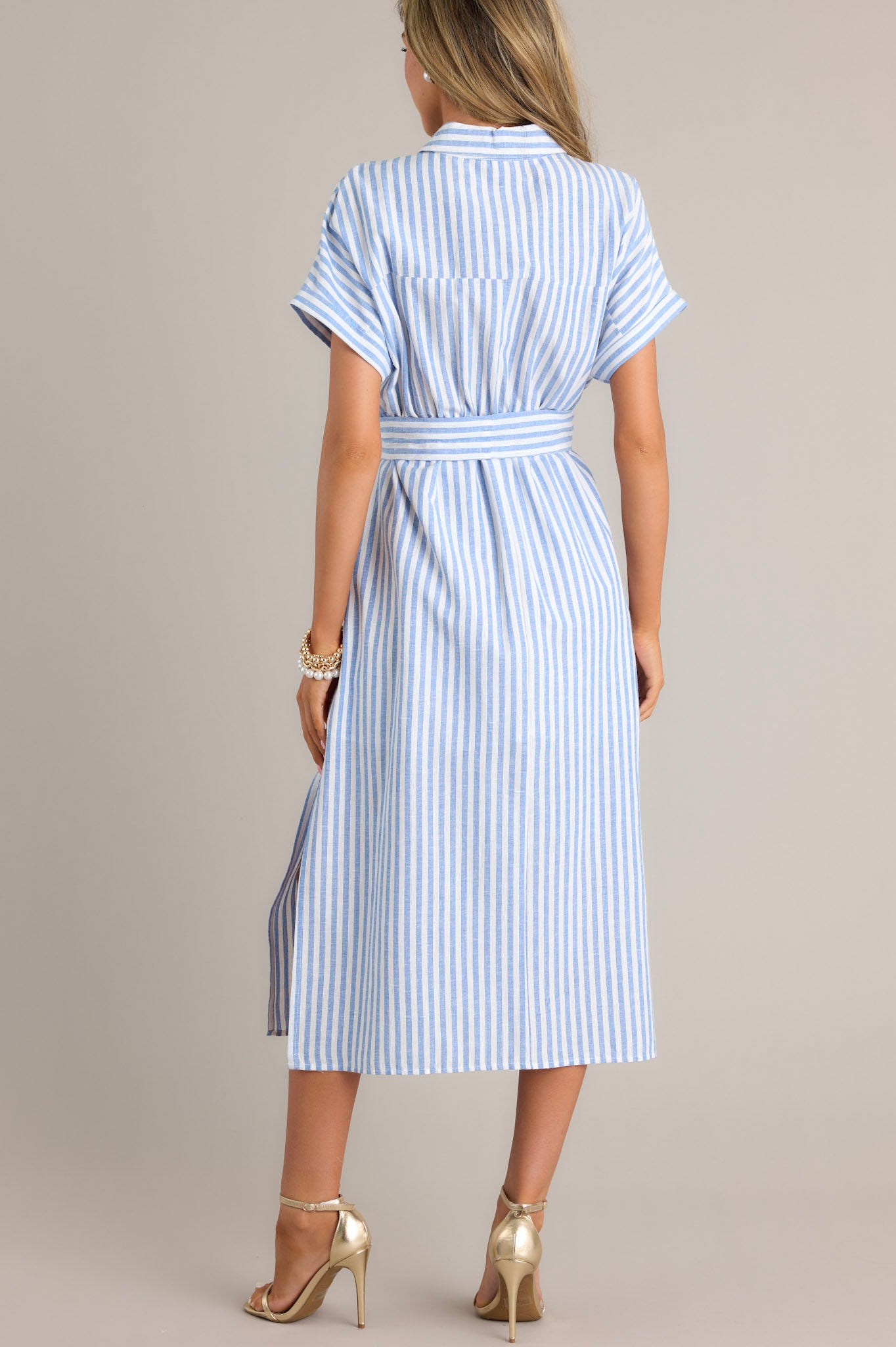 Back view of this dress that features a collared neckline, a full button front, a fitted waist, a self-tie waist belt, functional pockets, two side slits, and folded short sleeves.