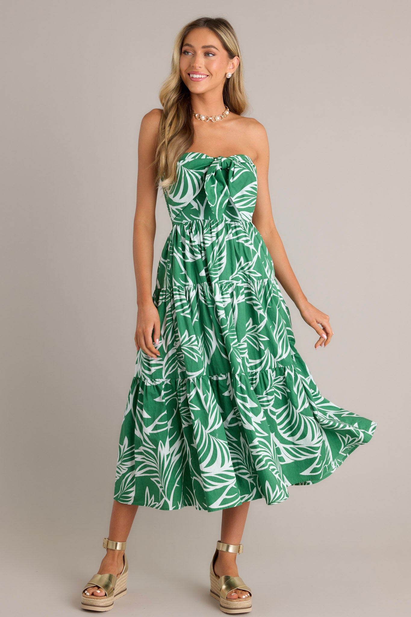 Front angled view of a green midi dress featuring a strapless neckline, boning in the bodice, a self-tie feature, a fully smocked insert in the back, and a tiered design