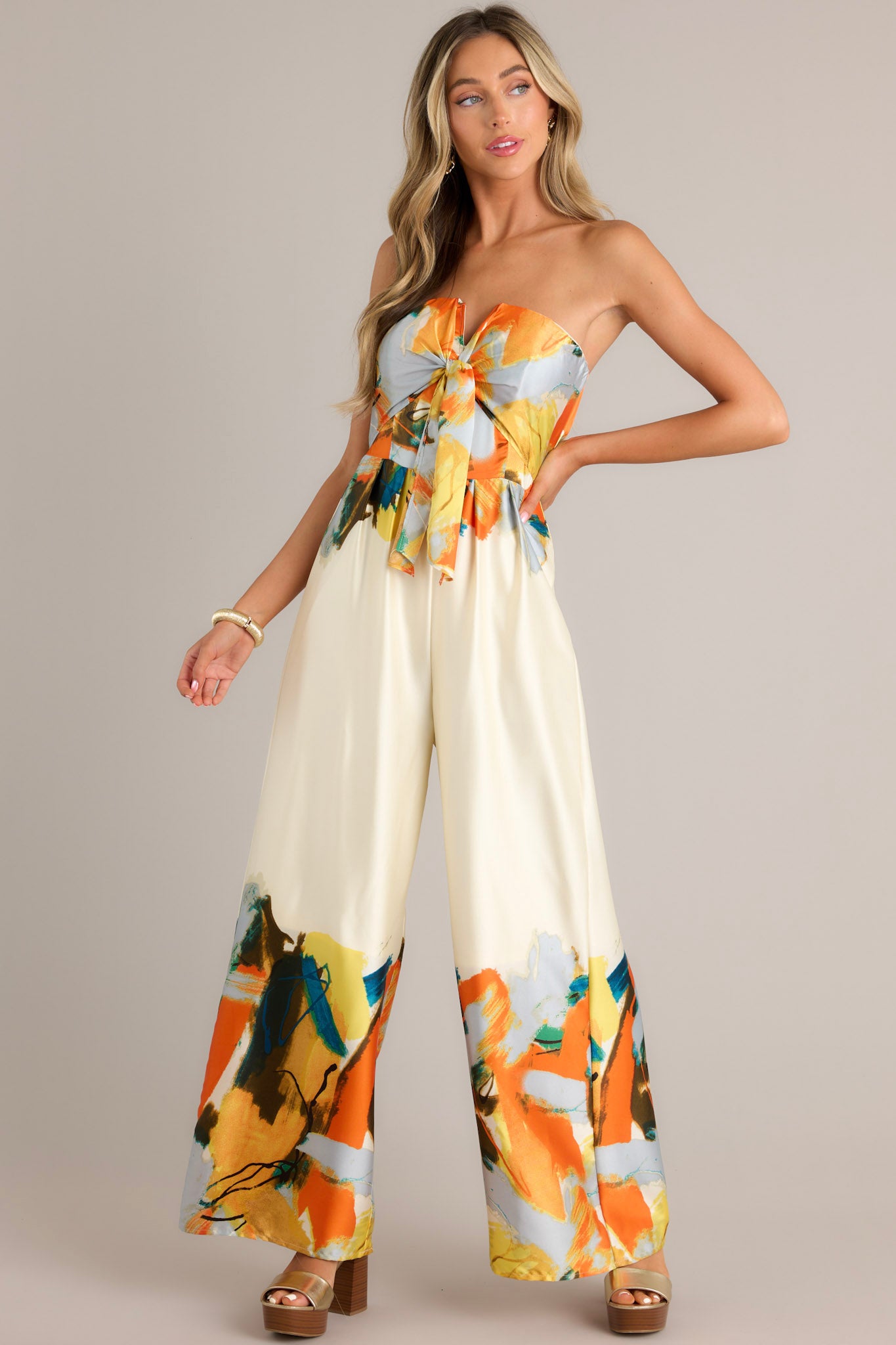 Front view of this strapless ivory multi jumpsuit with a tie front detail at the bust, wide leg pants, and abstract print of orange, blue, yellow, and green on the bust, waist, and bottom of the pants.