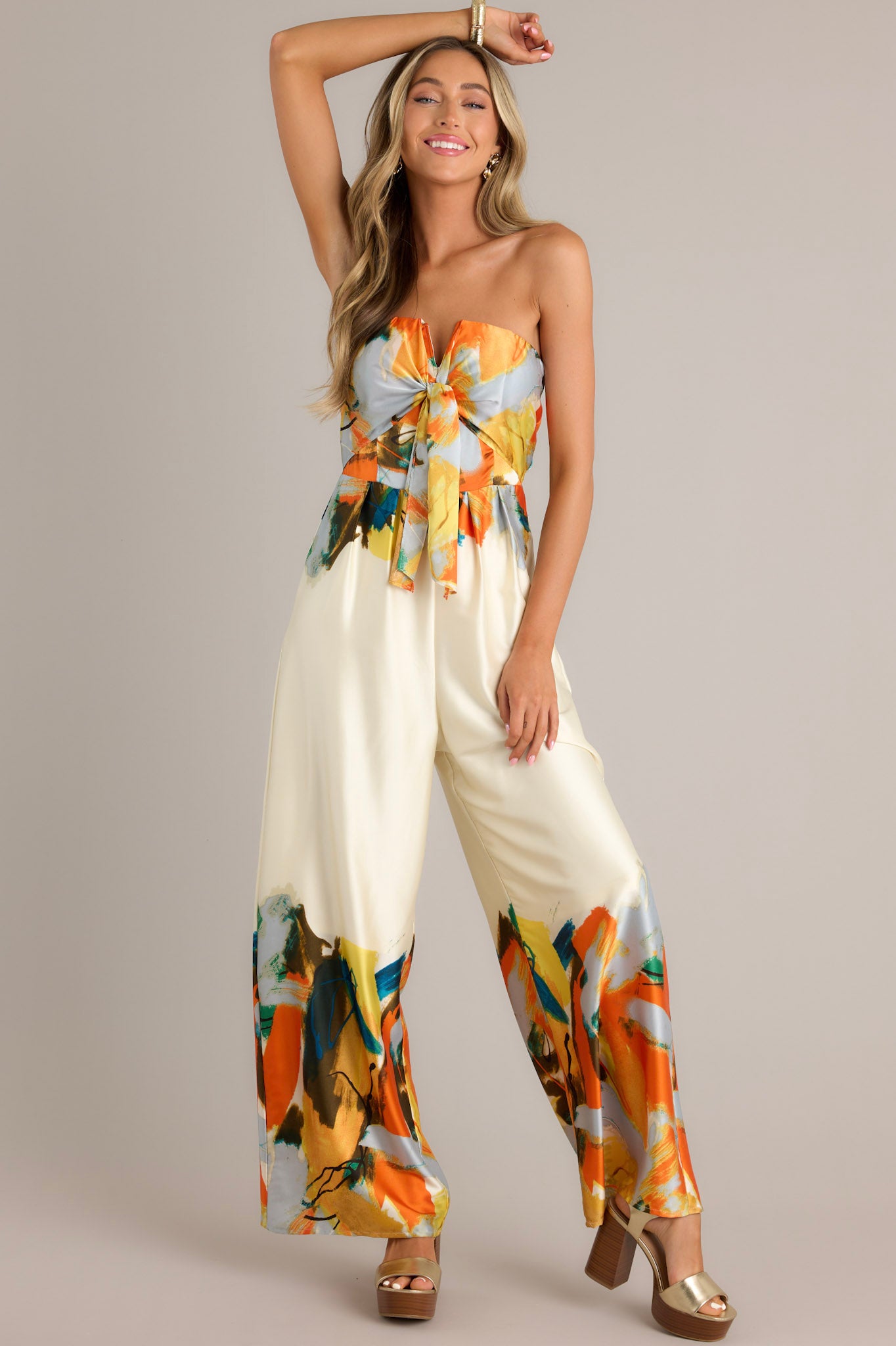 Strapless ivory multi jumpsuit with a tie front detail at the bust, wide leg pants, and abstract print of orange, blue, yellow, and green on the bust, waist, and bottom of the pants.