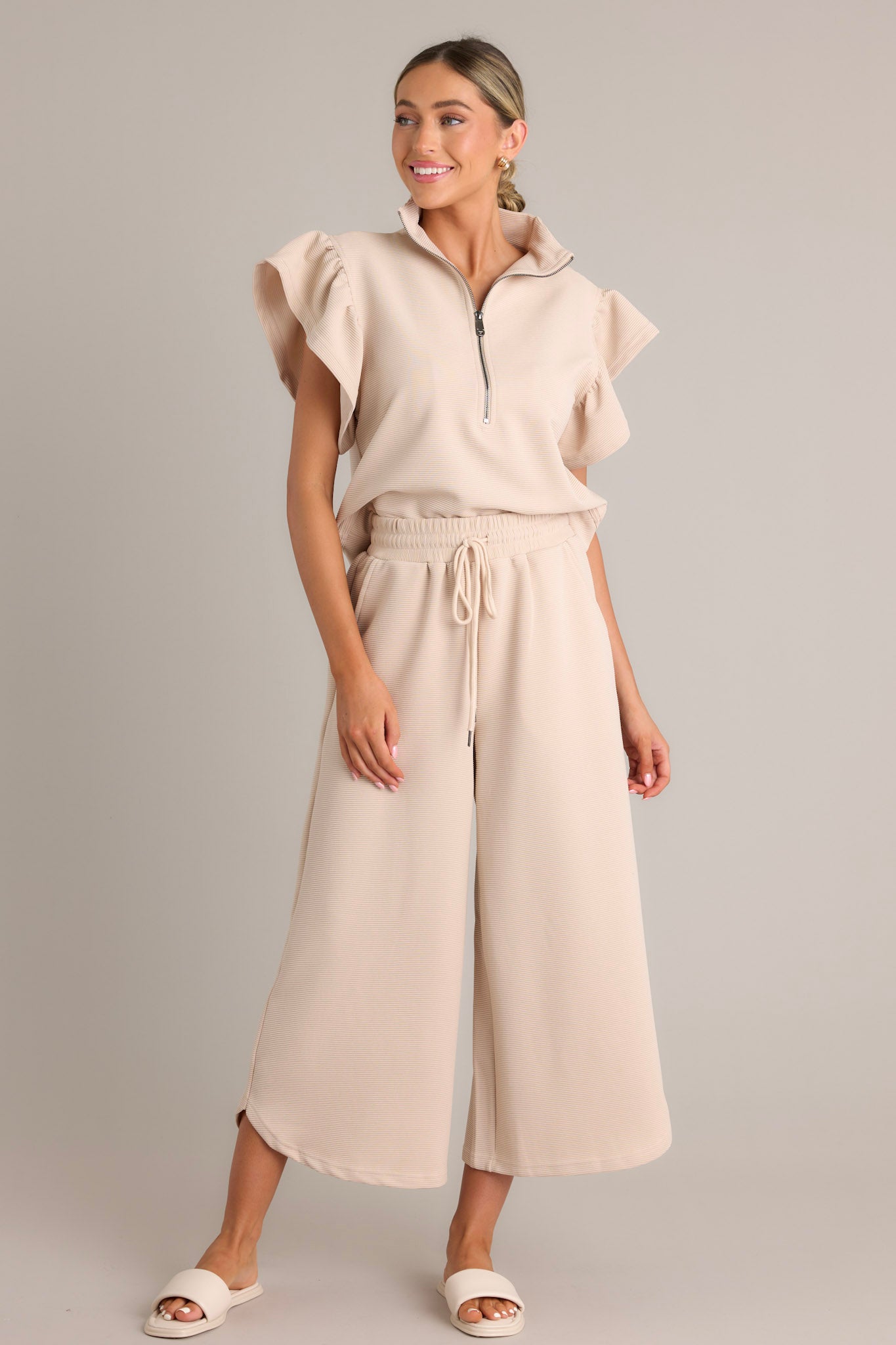 Full length view of a tan top featuring a functional zip-up neckline, ribbed fabric, wide ruffled sleeves, and a thick hemline.