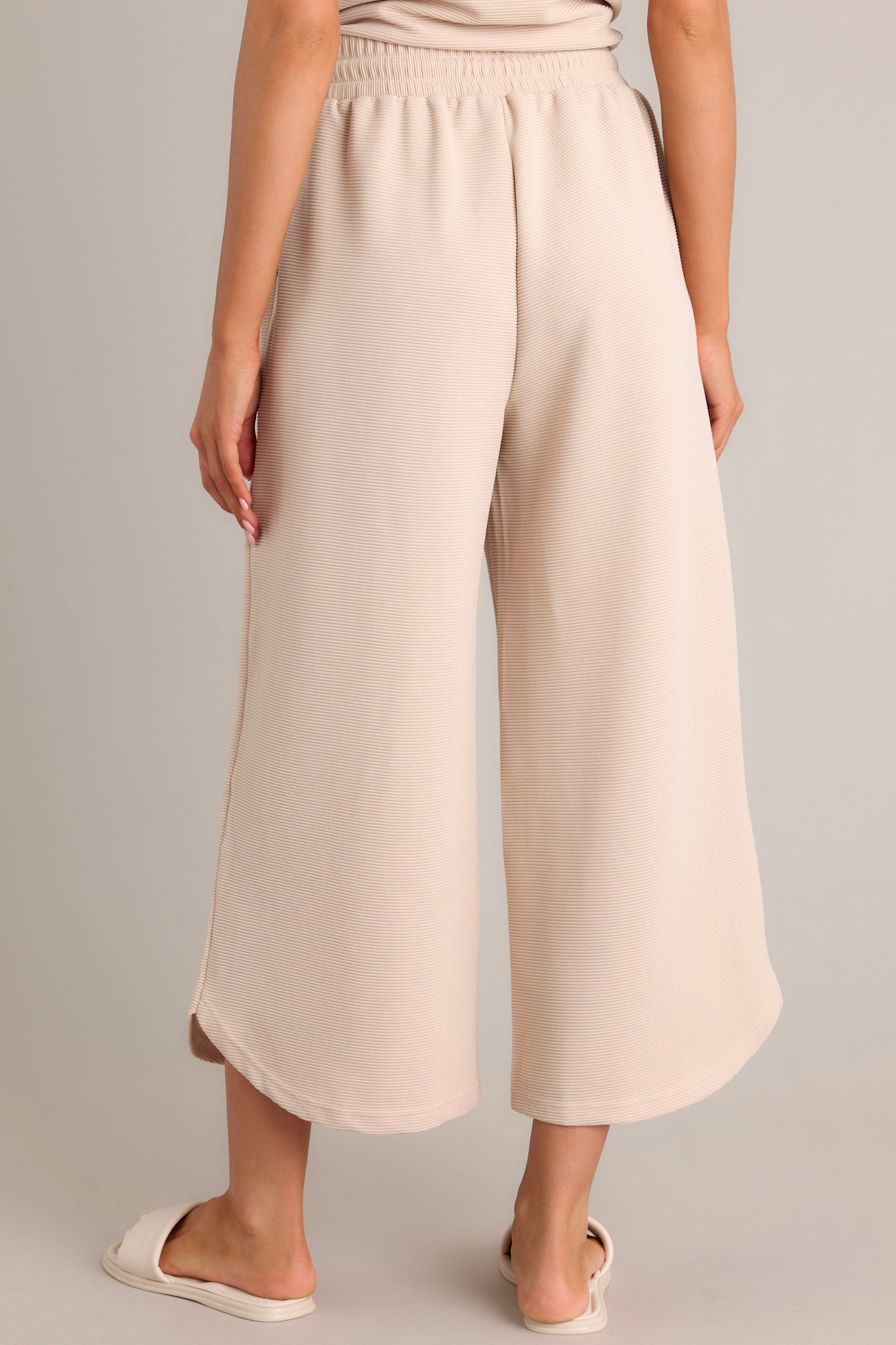 Back view of tan pants showcasing the high waisted design, elastic waistband, self-tie drawstring, ribbed texture, wide leg, and scooped hemline.