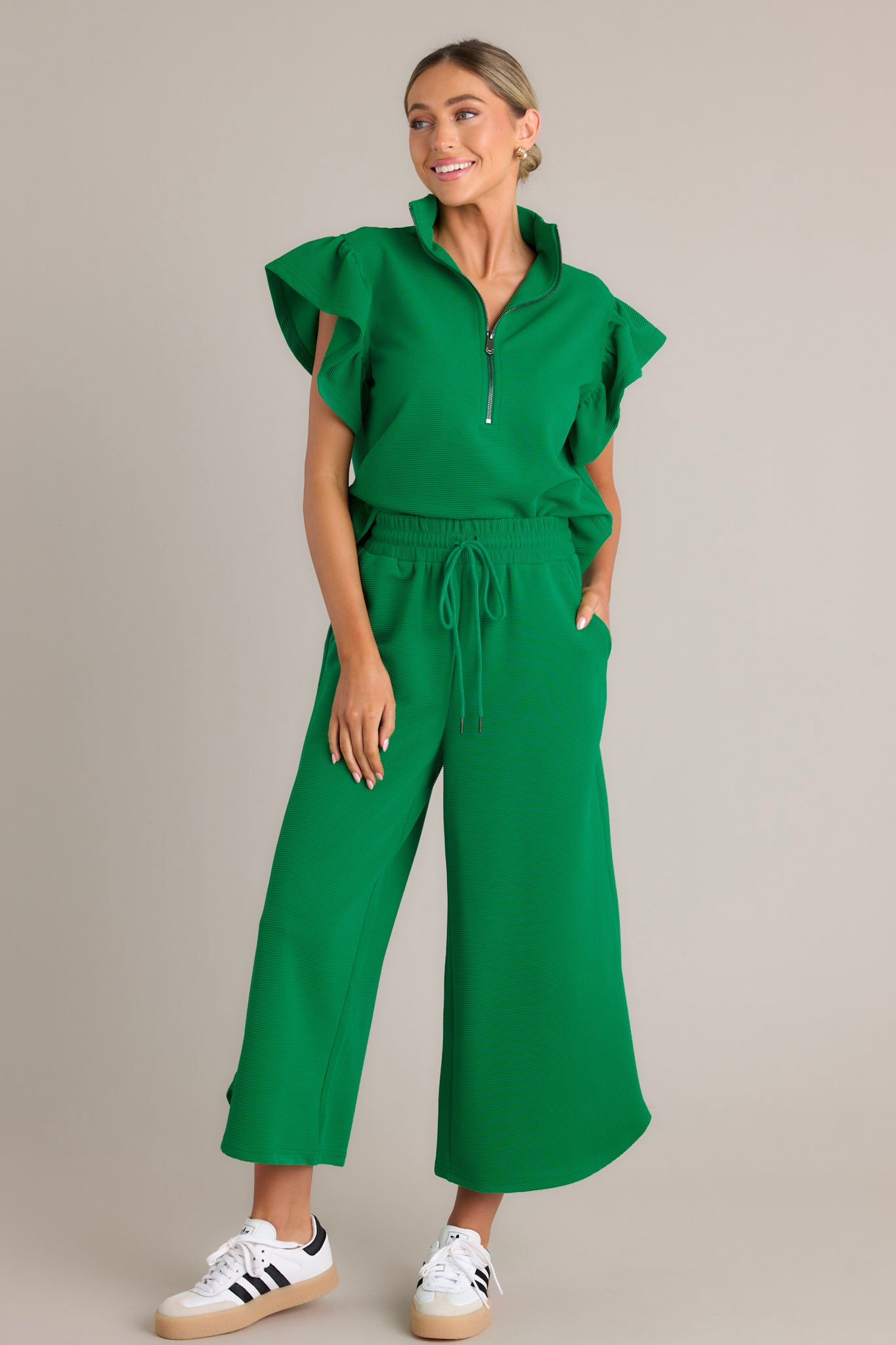 Front angled view of a green top featuring a functional zip up neckline, ribbed fabric, wide ruffled sleeves, and a thick hemline