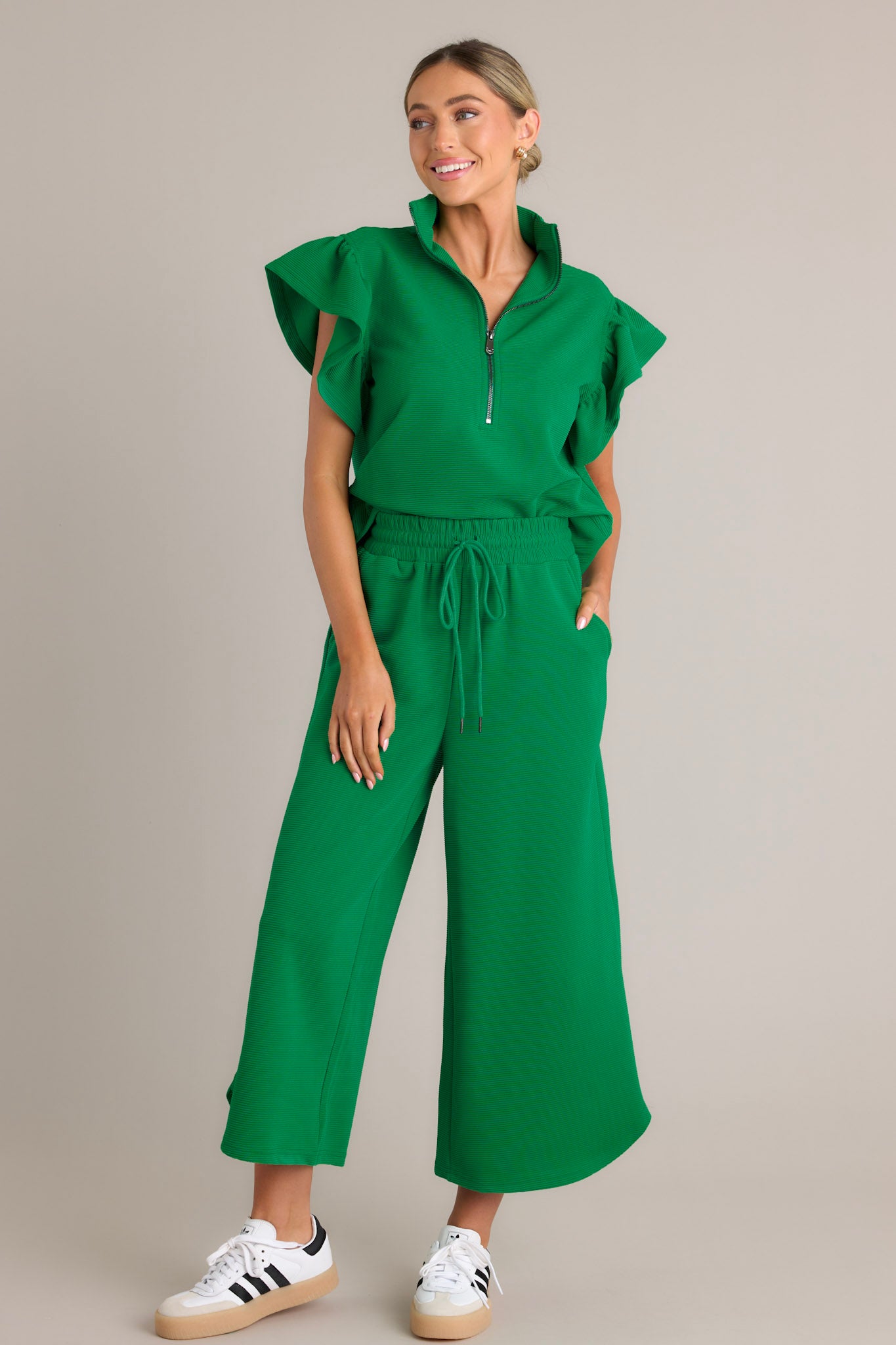 Full length view of green pants with a high waisted design, an elastic waistband, a self-tie drawstring, functional hip pockets, ribbed texture, wide leg, and a scooped hemline
