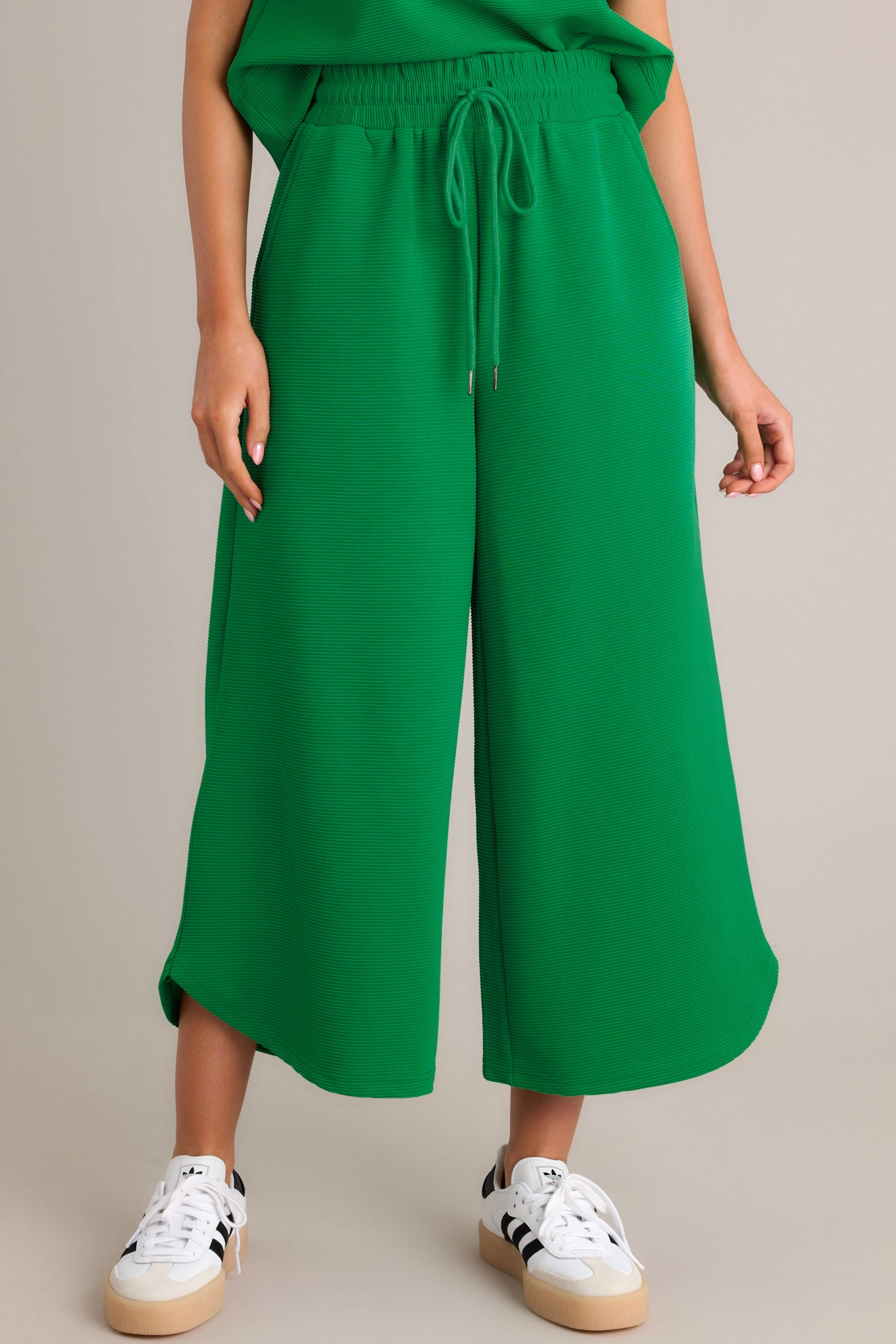 Front view of green pants featuring a high waisted design, an elastic waistband, a self-tie drawstring, functional hip pockets, a ribbed texture, a wide leg, and a scooped hemline.