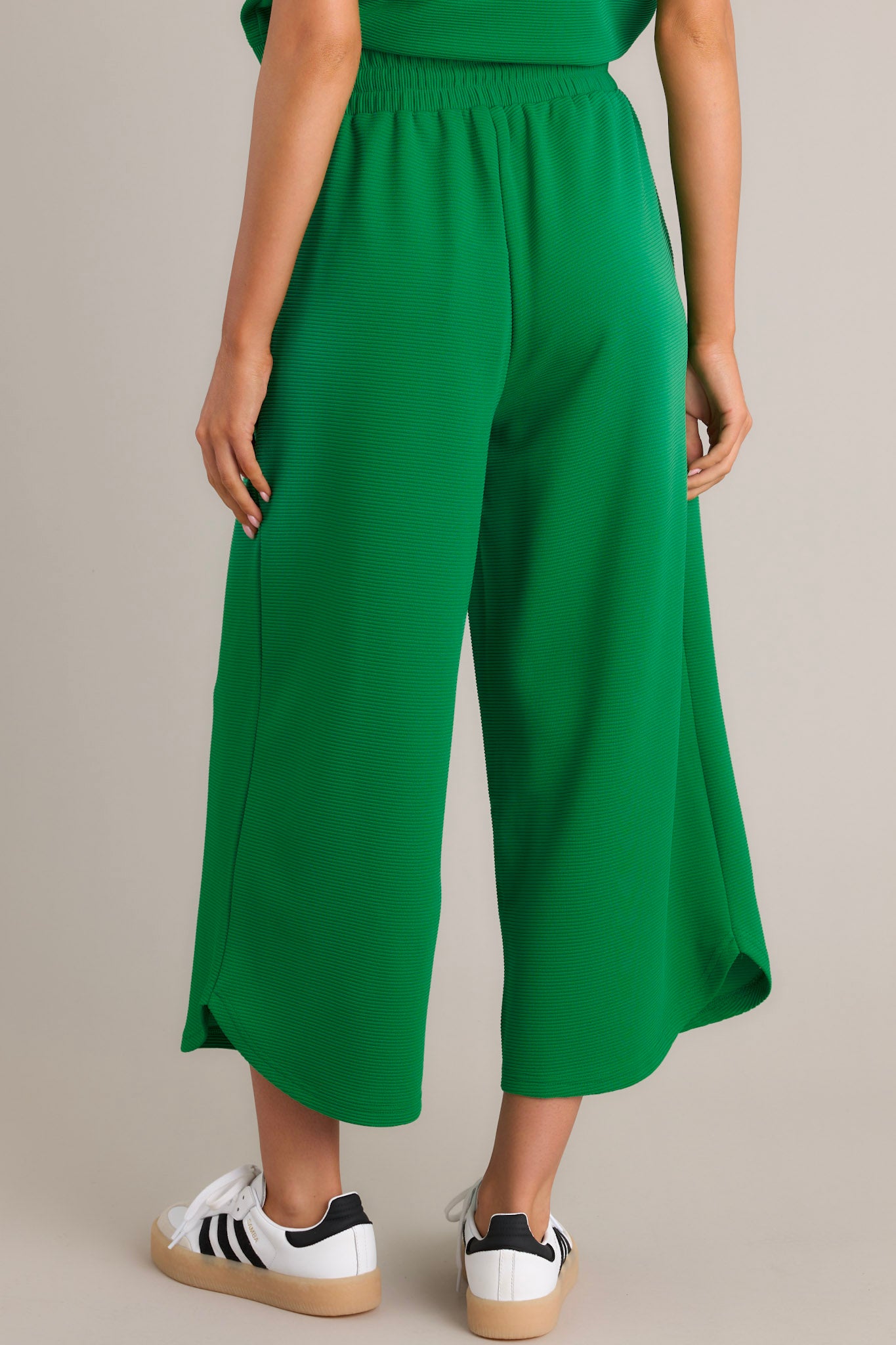 Back view of green pants highlighting the high waisted design, elastic waistband, ribbed texture, wide leg, and scooped hemline.