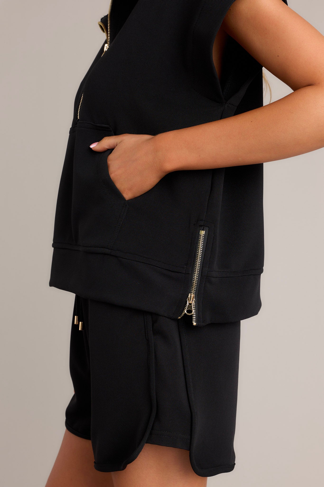 Side view of a black pullover showcasing the collared neckline, gold hardware, functional half zip design, kangaroo pocket, functional zippers on the sides, and short cap sleeves.
