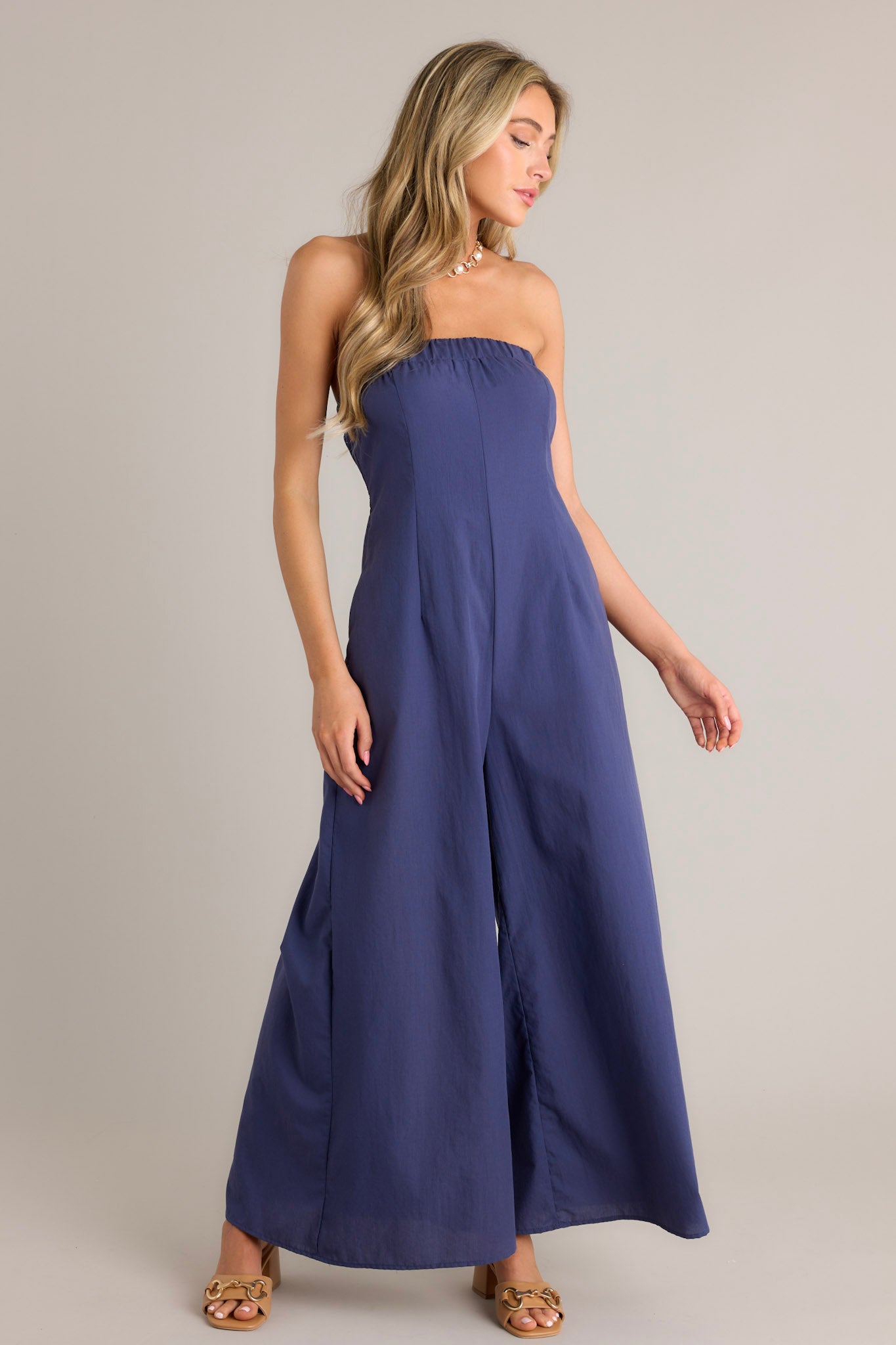 Full length view of a navy jumpsuit with an elastic strapless neckline, discrete side zipper, elastic cut-out in the back, functional hip pockets, and a wide leg