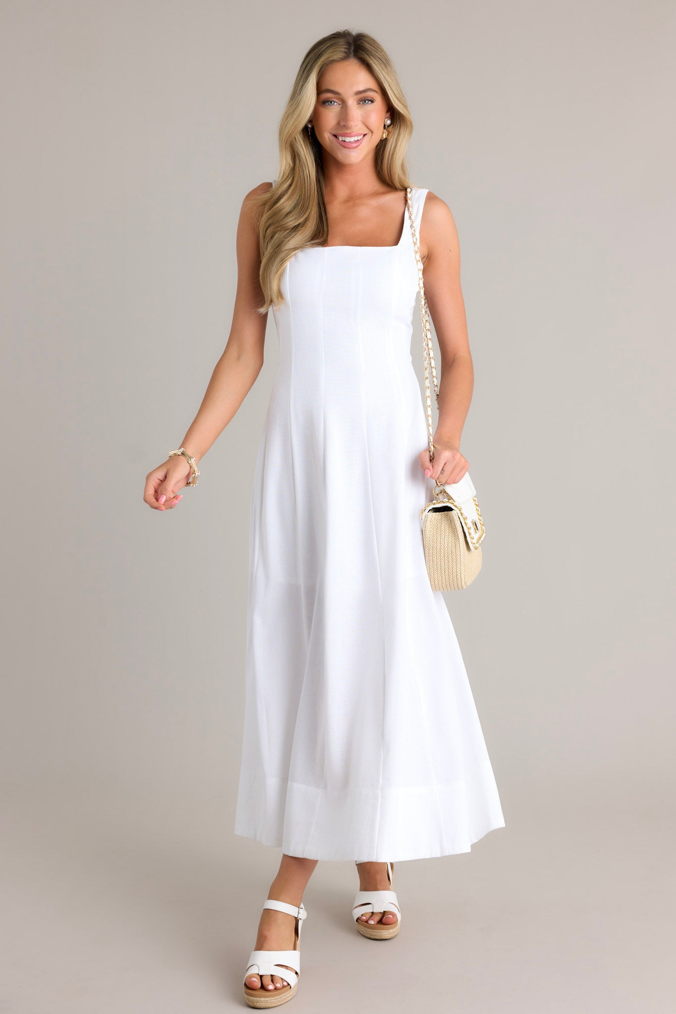 Front angled view of a white maxi dress featuring a square neckline, thick straps, a discrete back zipper, a flowing silhouette, and a thick hemline