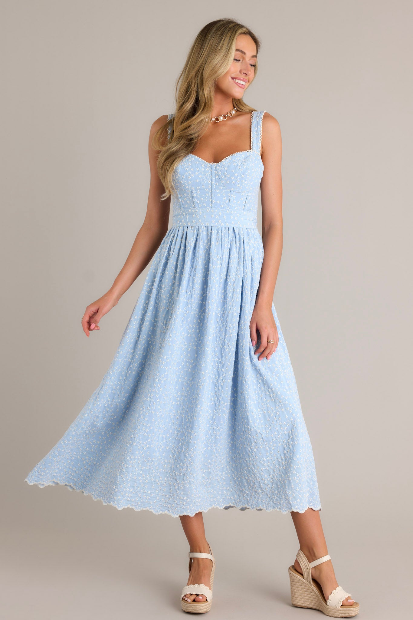 Full length view of a light blue midi dress with a sweetheart neckline, a smocked back insert, a zipper, thick adjustable straps, boning in the bodice, a self-tie waist belt, and embroidery throughout