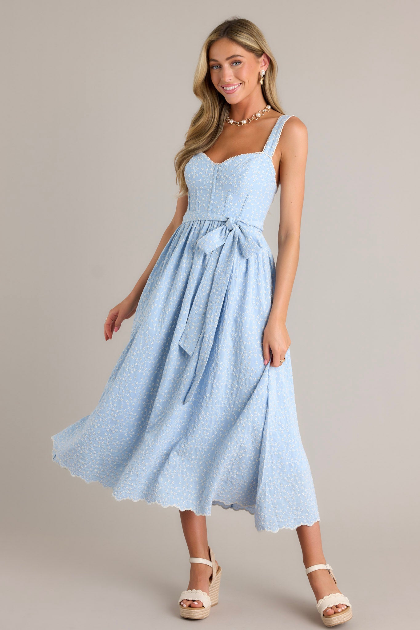 Front angled view of a light blue midi dress featuring a sweetheart neckline, a smocked back insert, a zipper, thick adjustable straps, boning in the bodice, a self-tie waist belt, and embroidery throughout