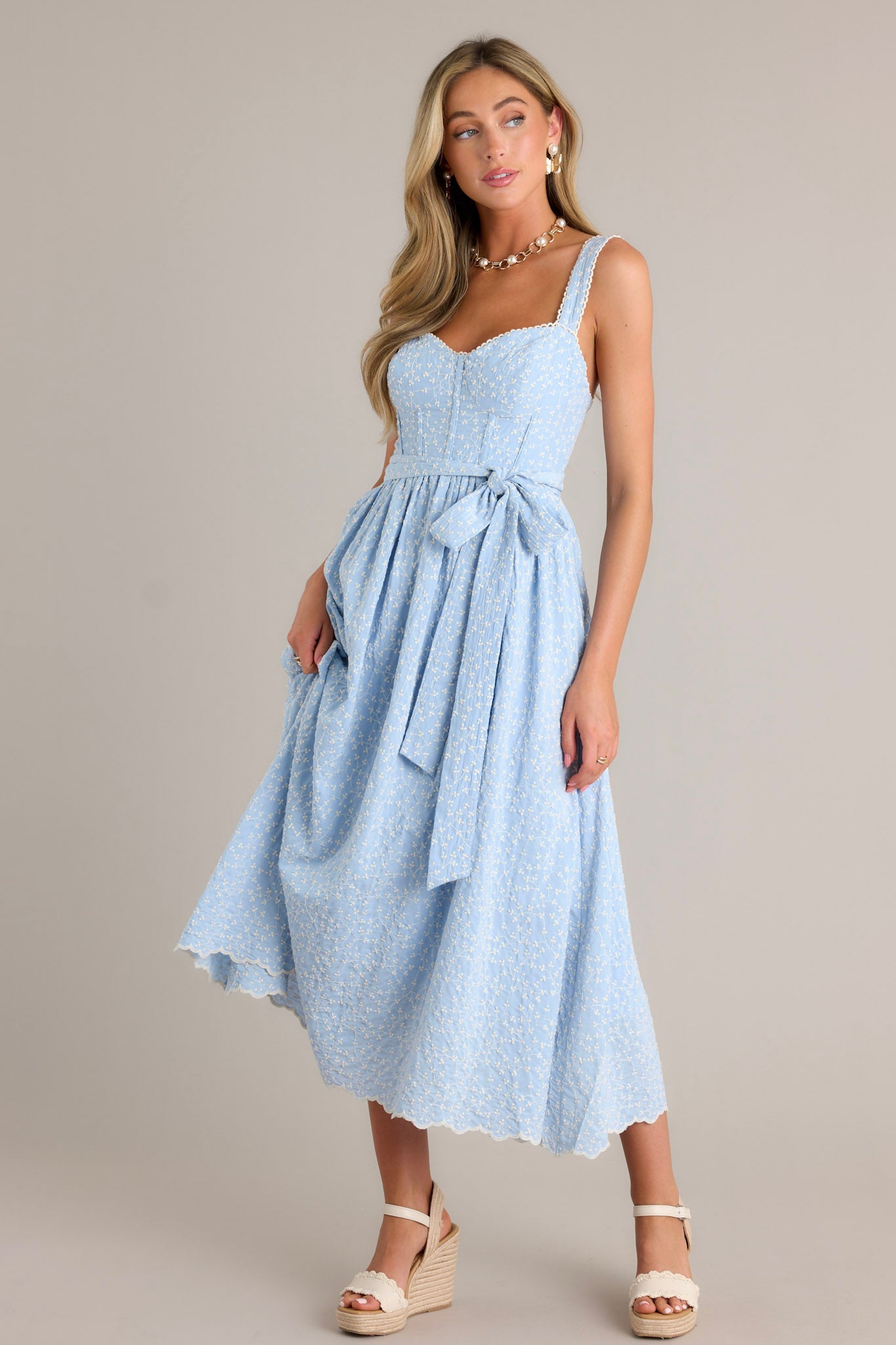 Front view of a light blue midi dress featuring a sweetheart neckline, thick adjustable straps, boning in the bodice, a self-tie waist belt, and embroidery throughout.