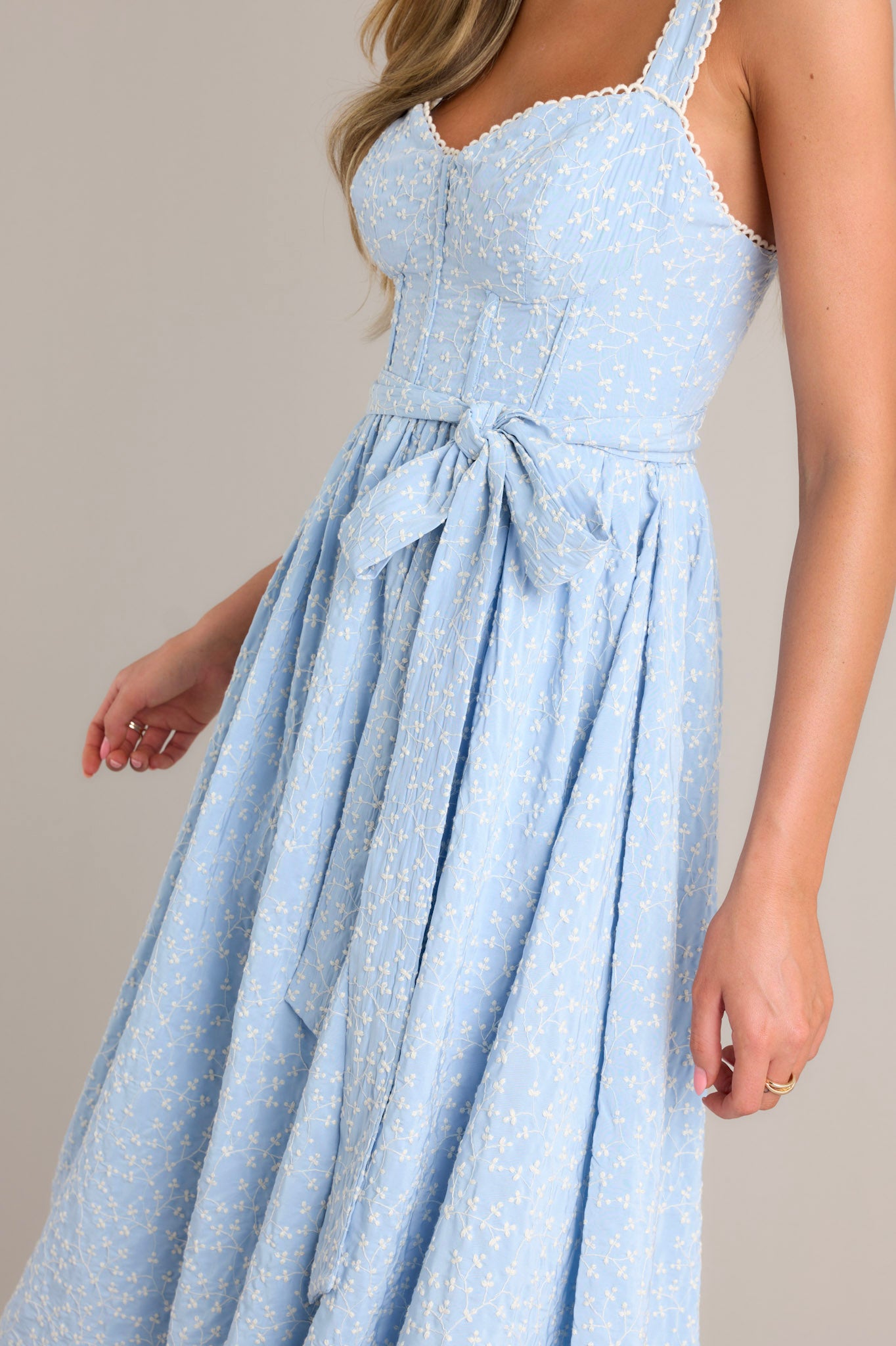 Side view of a light blue midi dress showcasing the sweetheart neckline, thick adjustable straps, boning in the bodice, self-tie waist belt, and embroidery throughout.