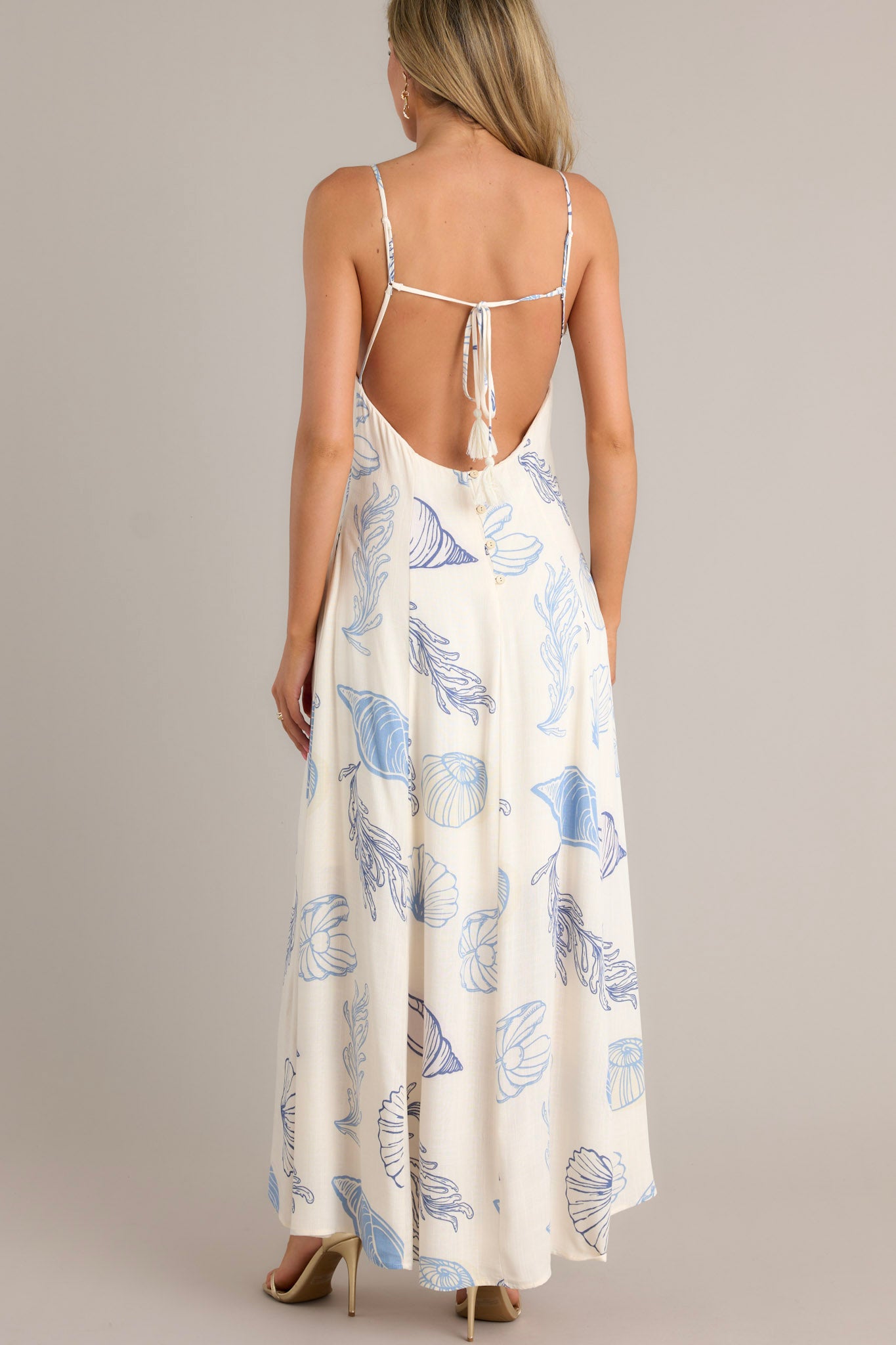 Back view of a blue maxi dress highlighting the low back with functional buttons, thin adjustable straps with a self-tie feature, and flowing silhouette.